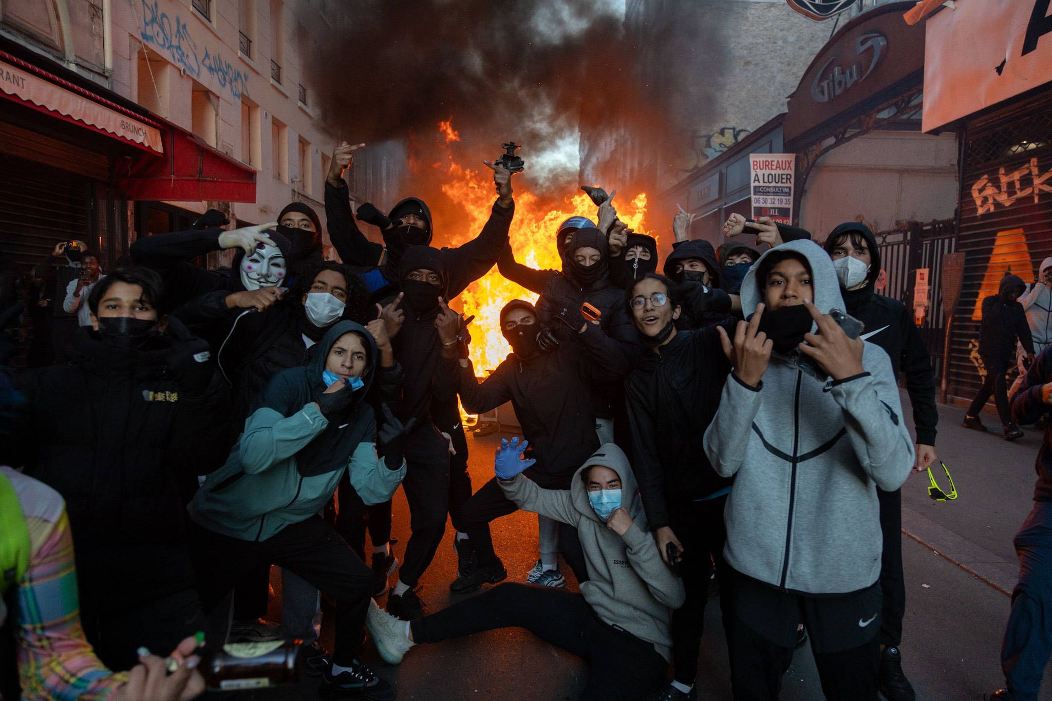 Gruppe junger Menschen mit Masken posiert vor brennender Barrikade bei Protest in Paris.