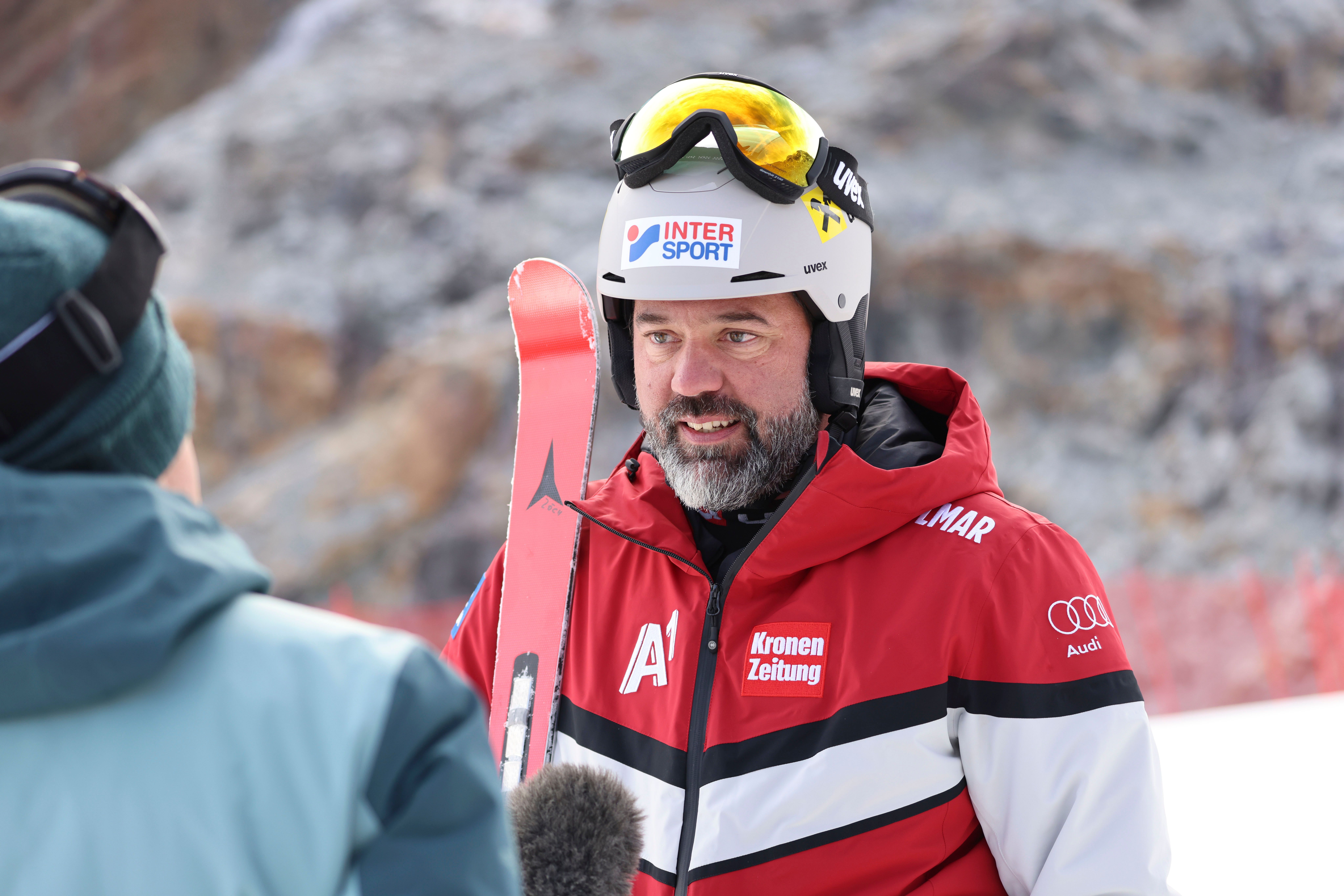 Ein Skitrainer im roten Anorak mit Helm und Skibrille spricht im Schnee mit einer Person.