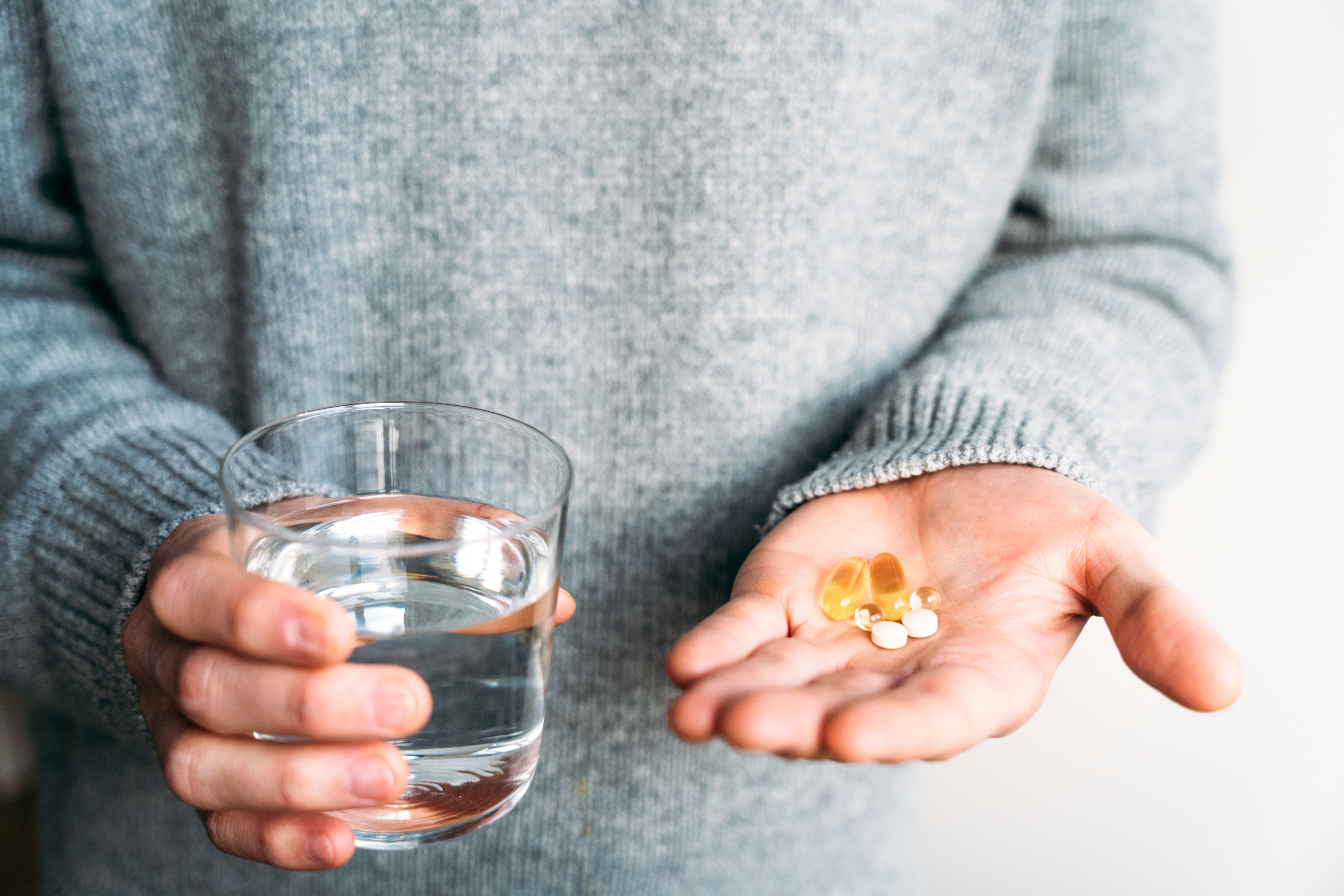 Person hält ein Glas Wasser in einer Hand und mehrere Nahrungsergänzungsmittel in der anderen.
