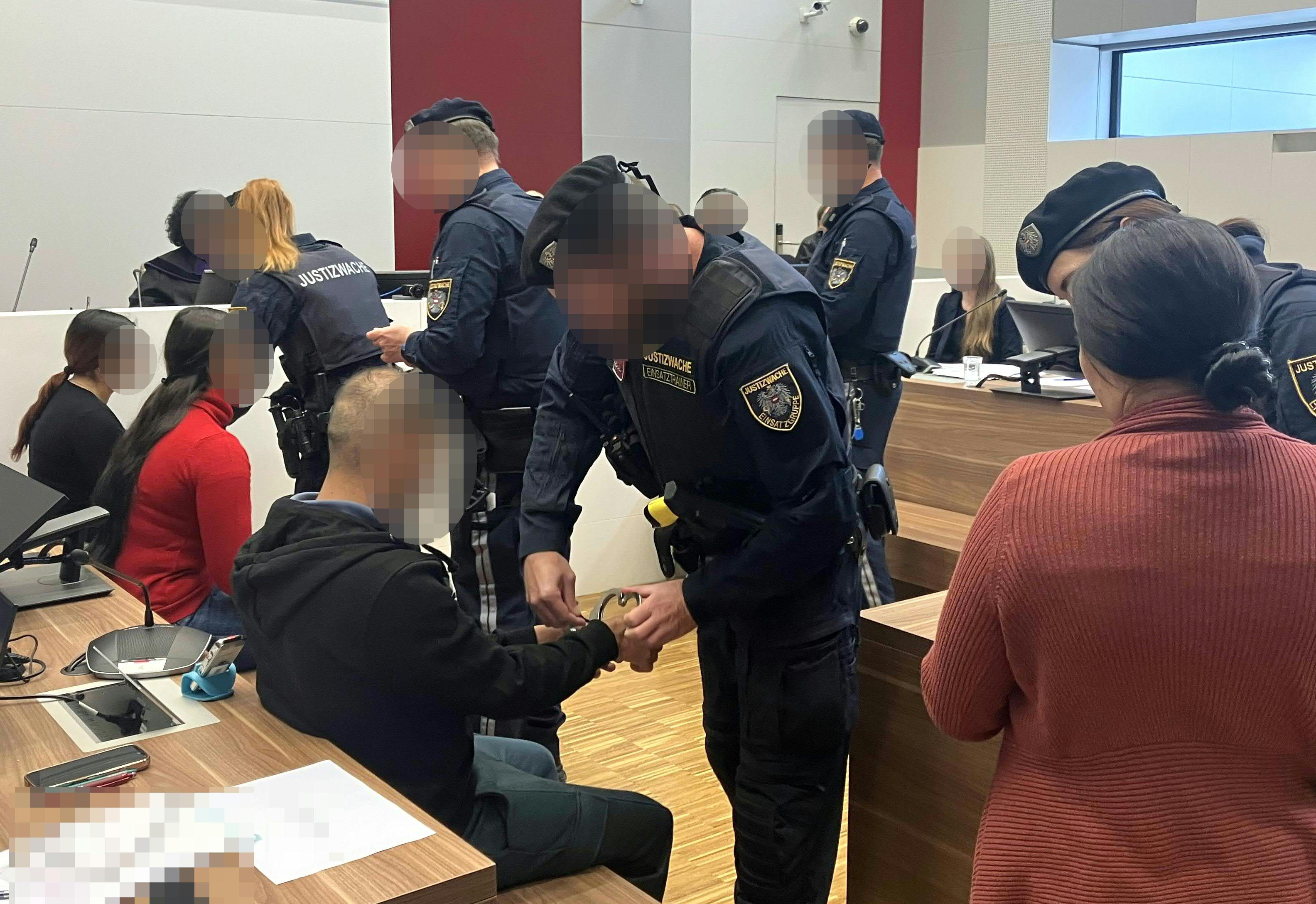 Mehrere Justizbeamte begleiten Angeklagte in Handschellen in einem Gerichtssaal.