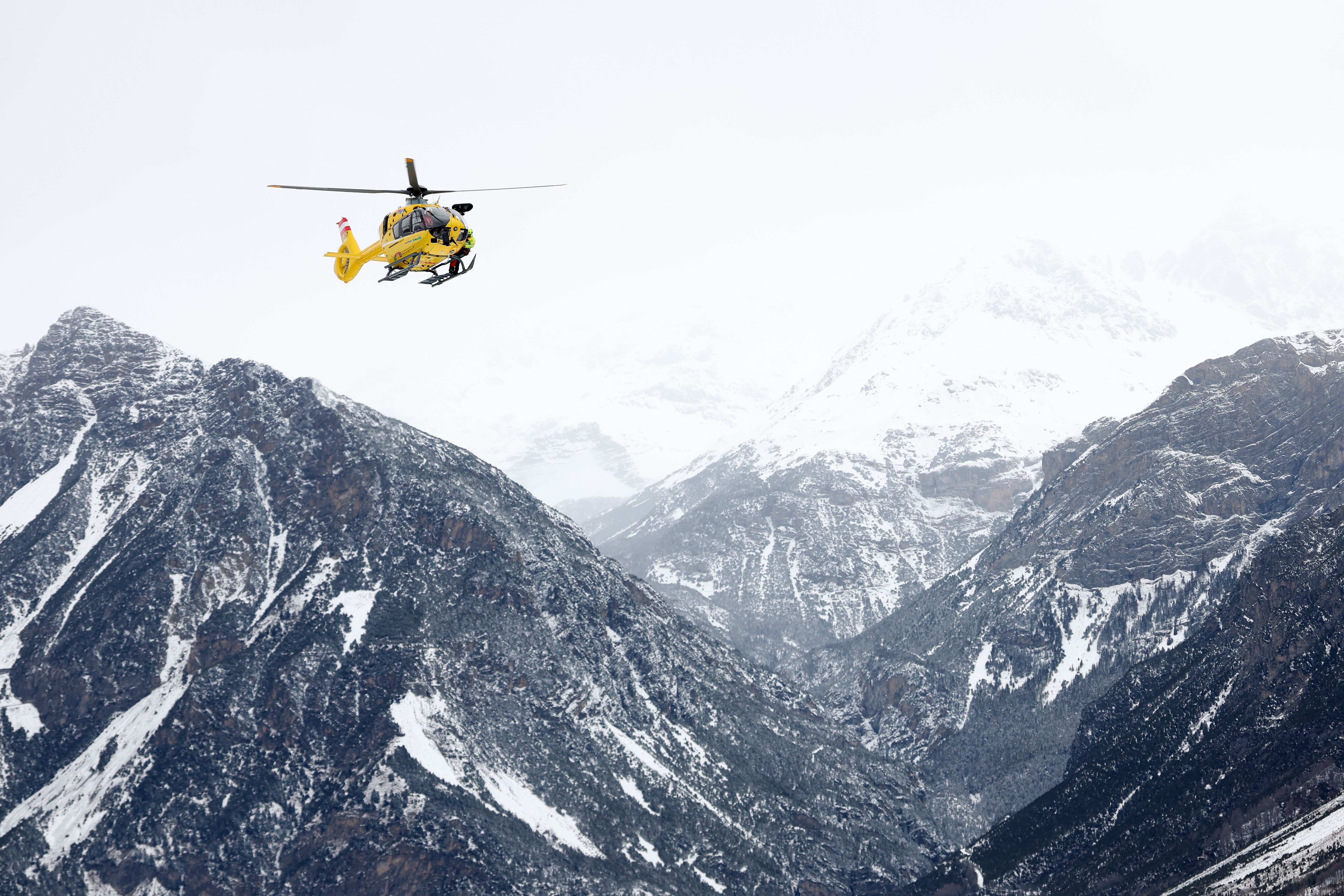 Ein gelber Rettungshubschrauber fliegt über verschneite Berge bei bewölktem Himmel.