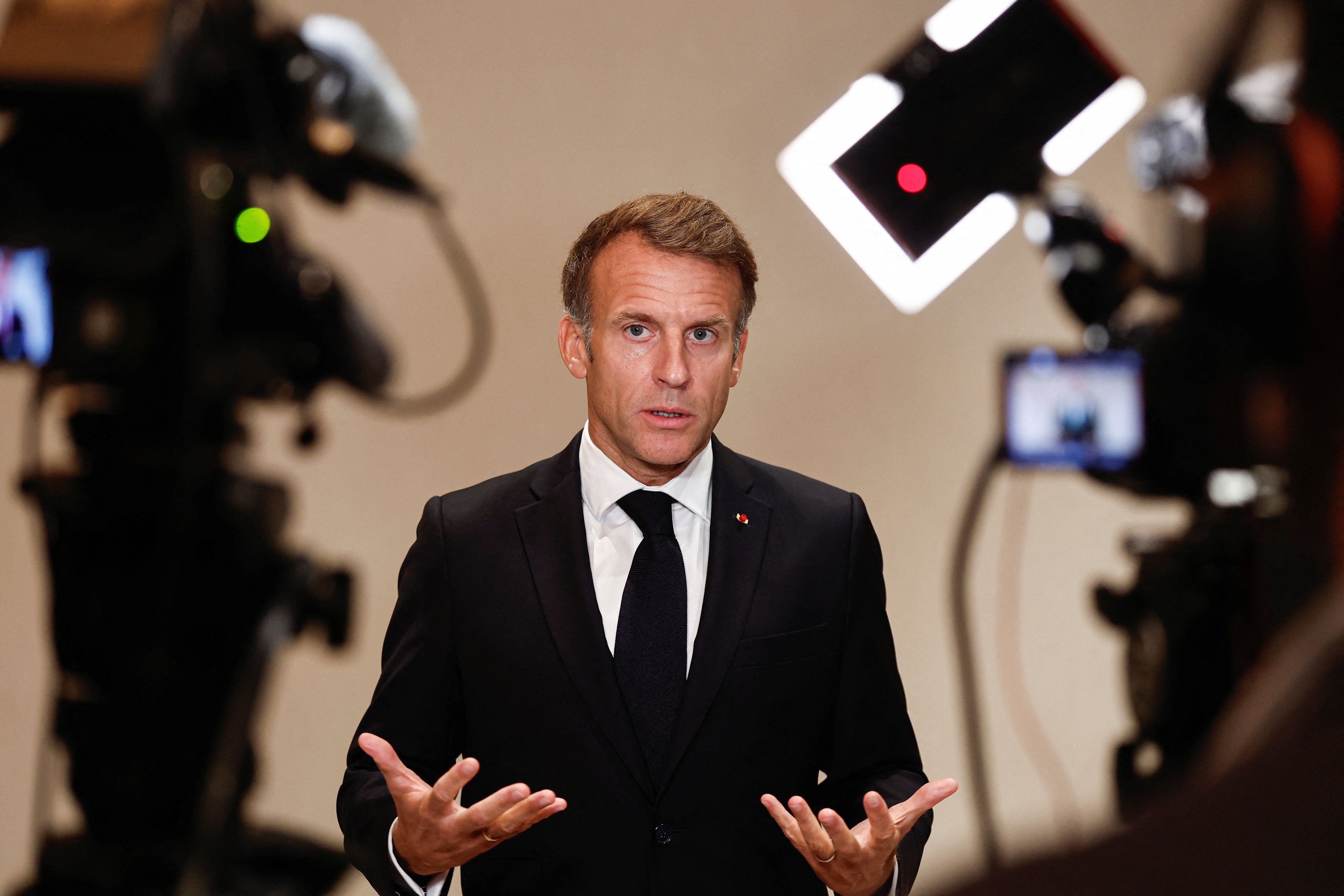 Ein Mann im Anzug spricht mit ernstem Gesicht zwischen Kameras vor neutralem Hintergrund.