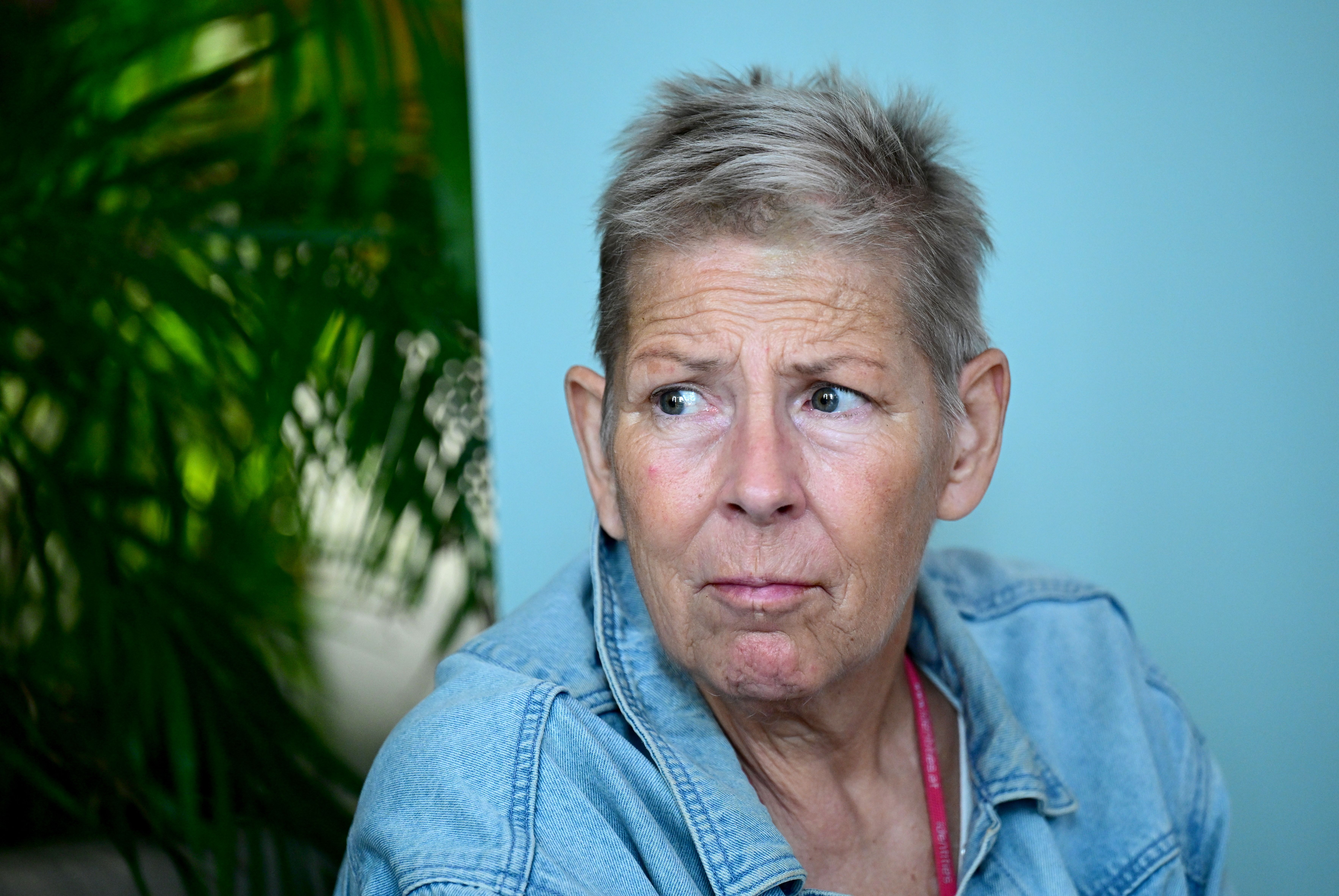 Eine nachdenklich blickende ältere Frau mit kurzen Haaren und Jeansjacke sitzt vor blauem Hintergrund.