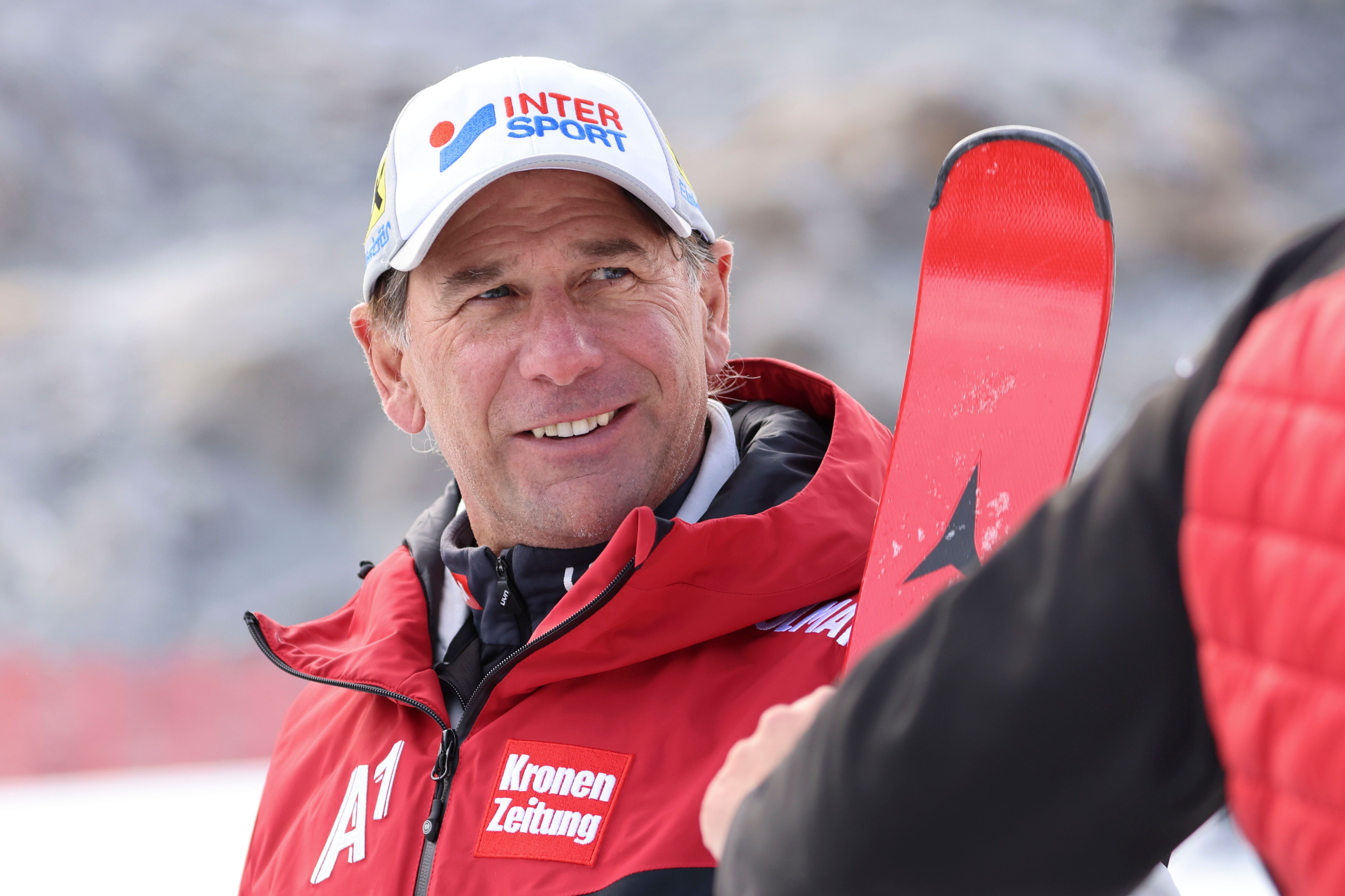 Ein Mann in roter Skijacke und Mütze steht im Schnee und hält Skier in der Hand.