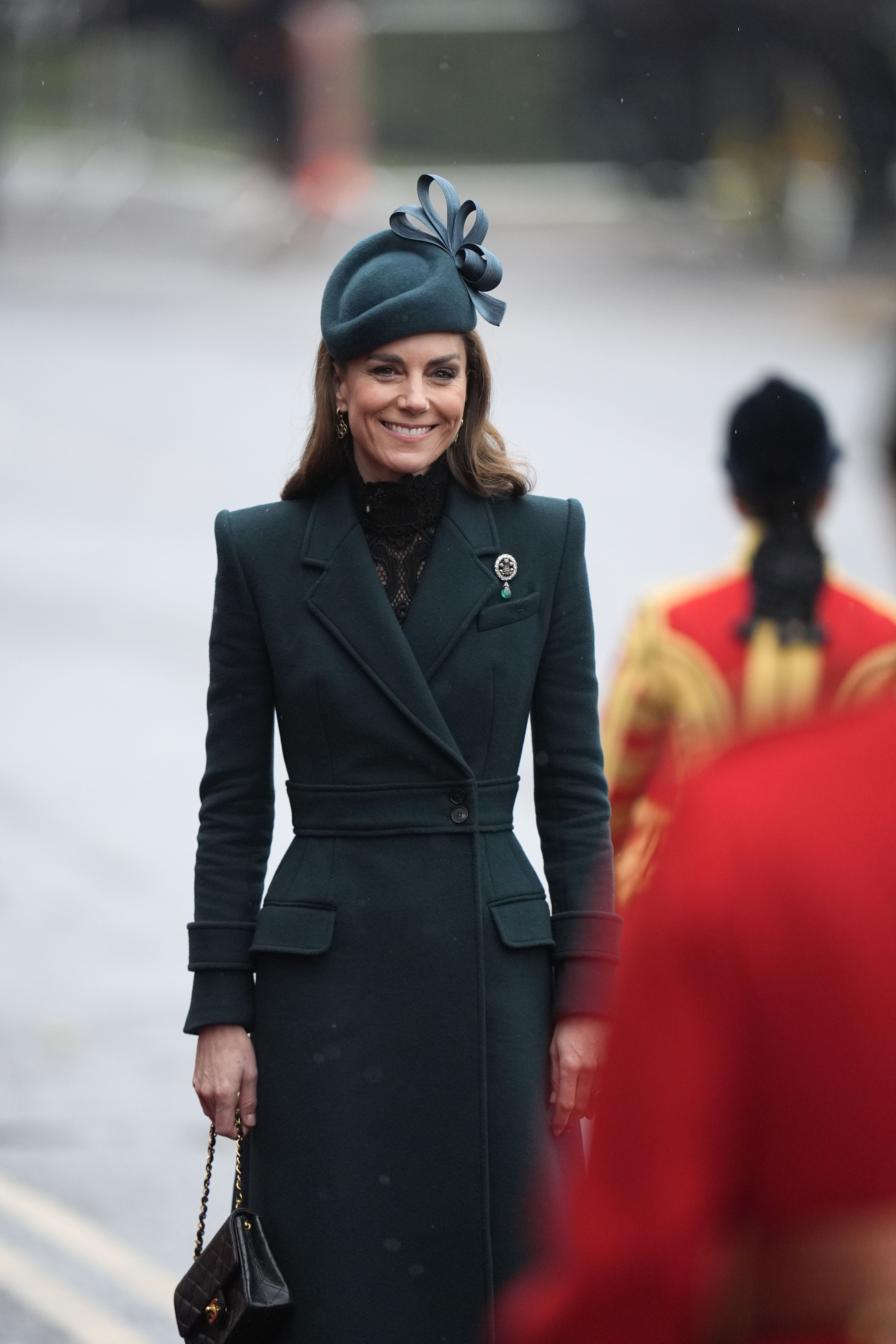 Eine elegant gekleidete Frau mit grünem Mantel und Hut lächelt bei einer Zeremonie im Freien.