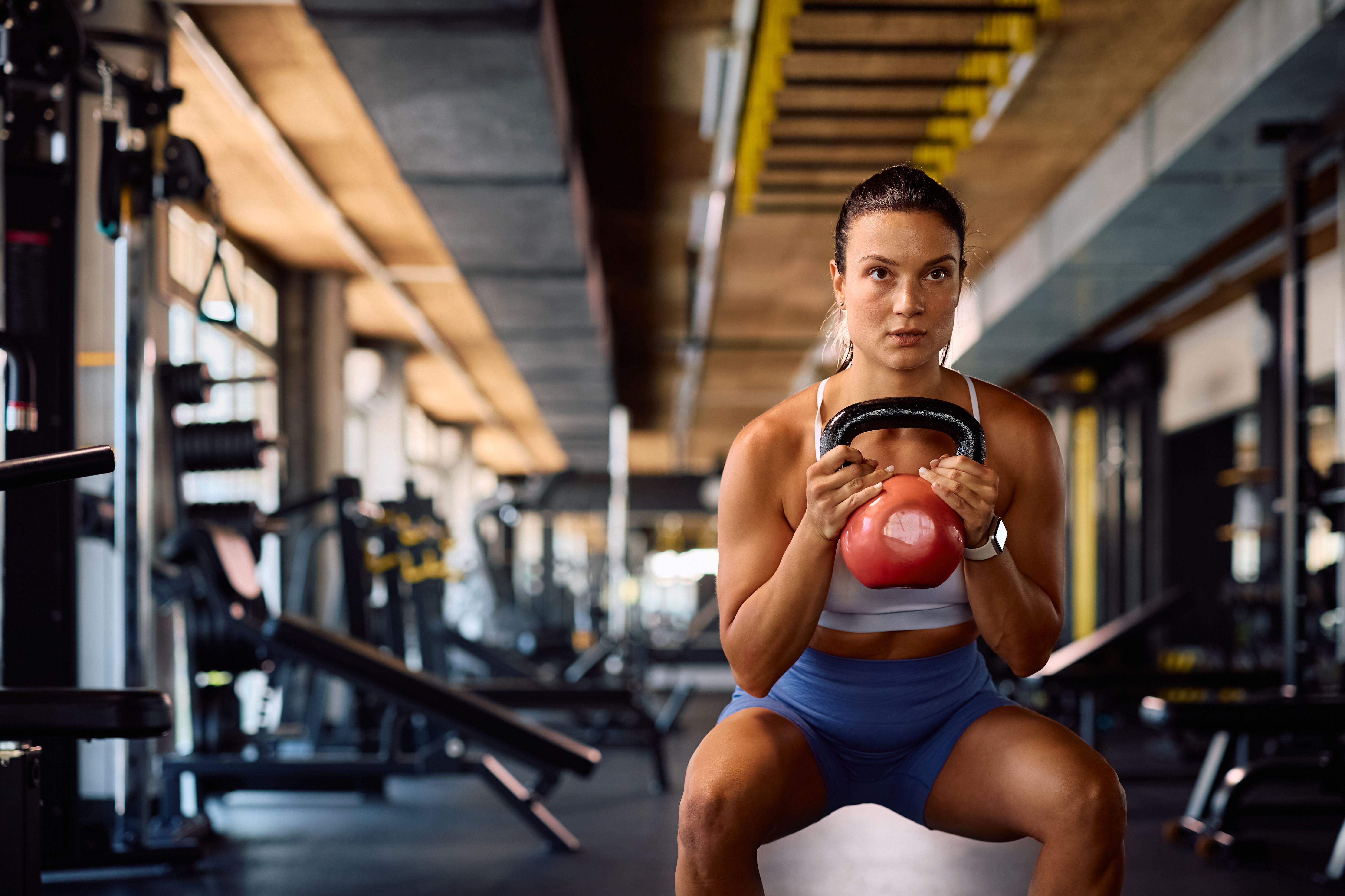 Junge Sportlerin macht Kniebeugen mit einer Kettlebell im Fitnessstudio.