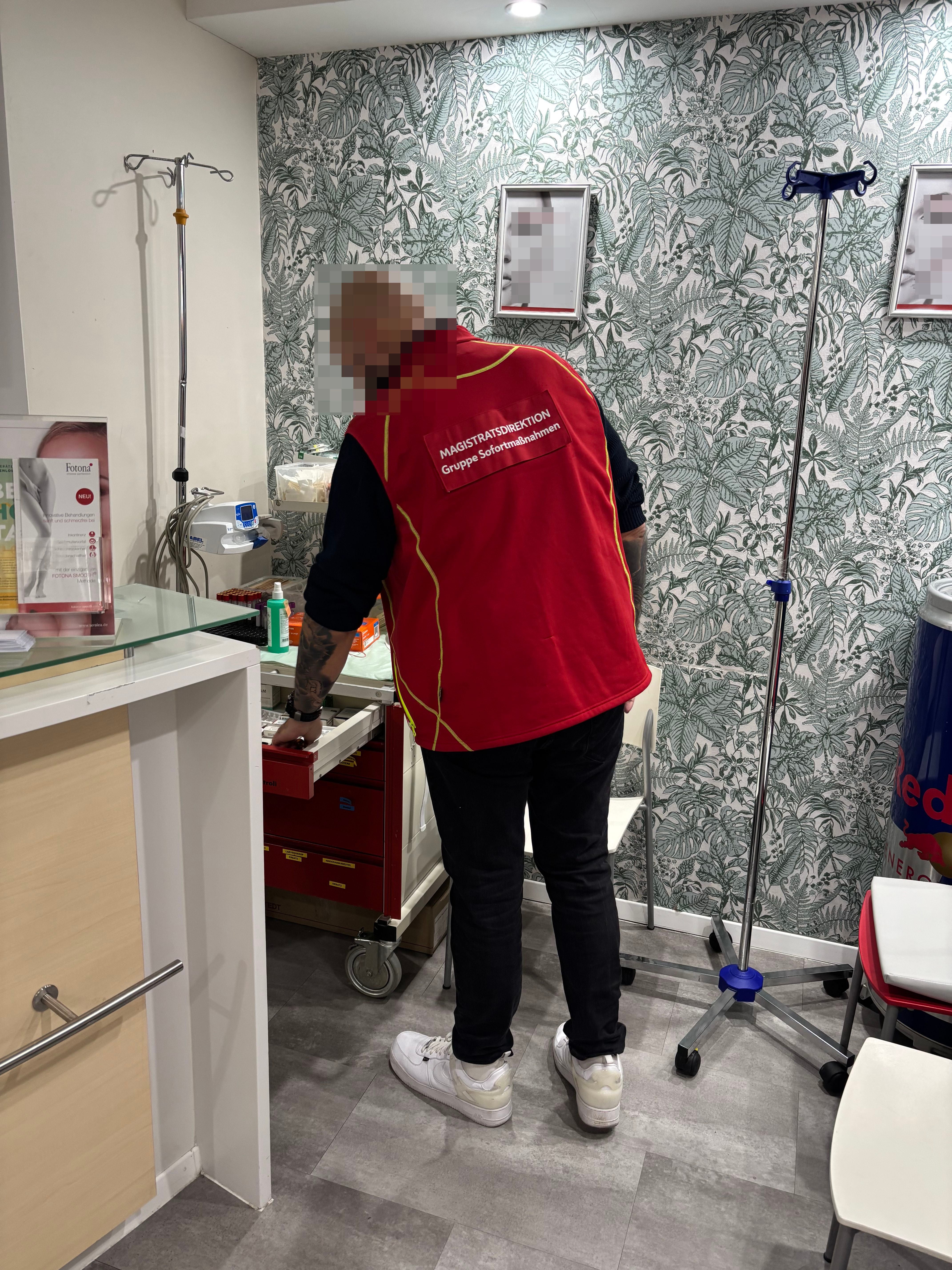 Person mit roter Weste arbeitet an einem offenen Medikamentenwagen in einem Behandlungsraum.