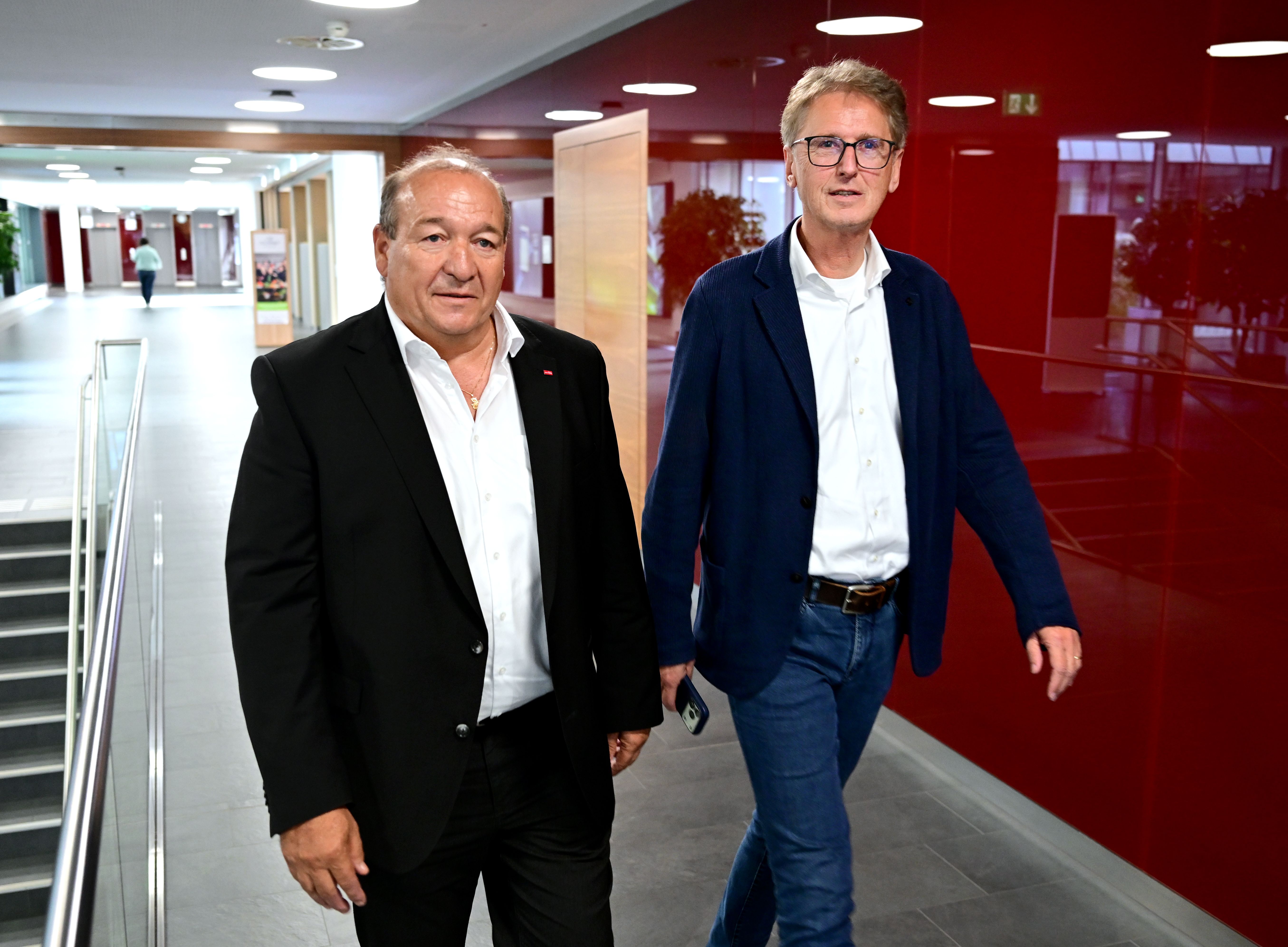 Zwei Männer in Business-Kleidung gehen durch einen modernen Flur mit roter Wand.