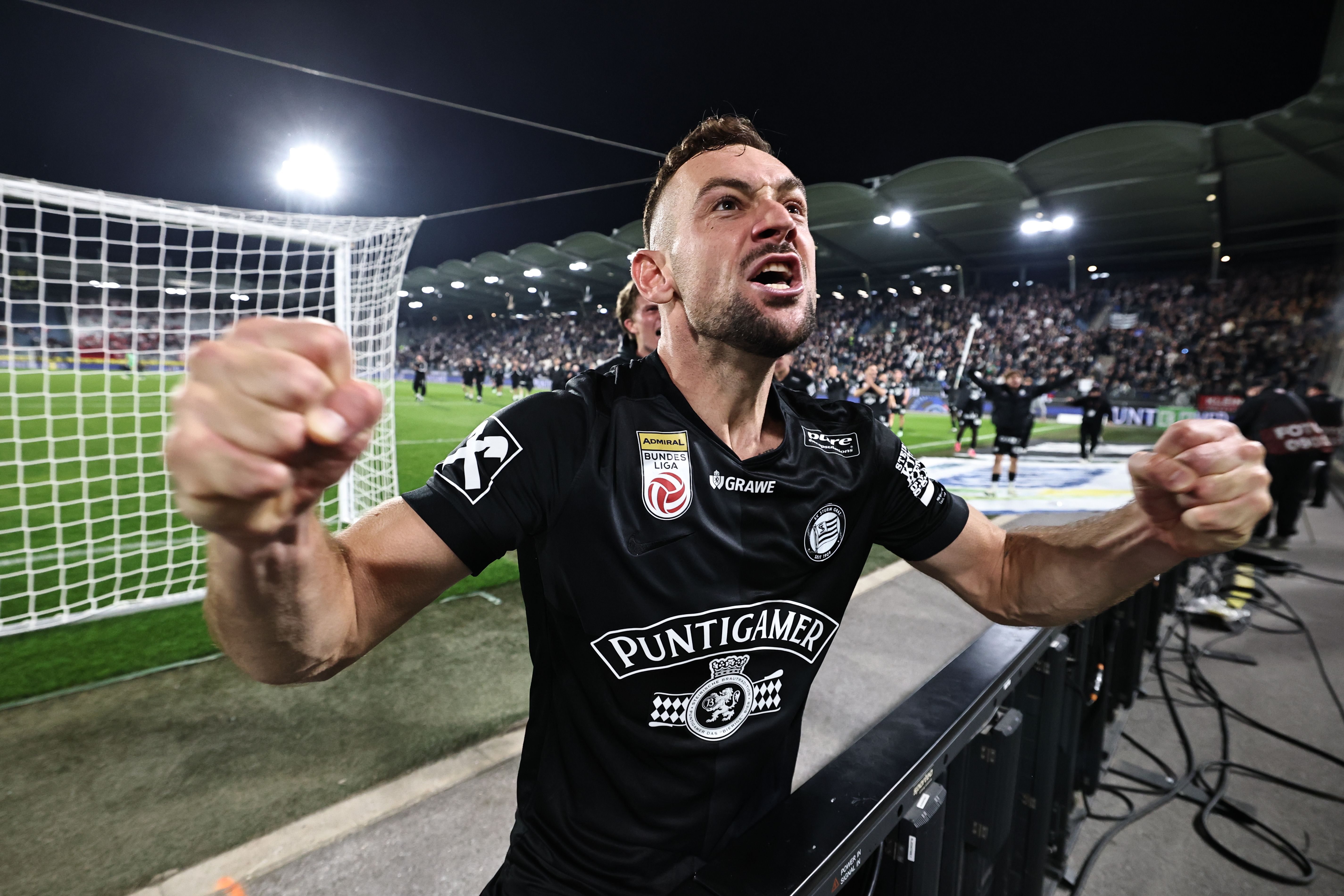 Ein Sturm-Graz-Spieler jubelt im schwarzen Trikot mit geballten Fäusten und lautem Schrei nah vor der Tribüne im Stadion.