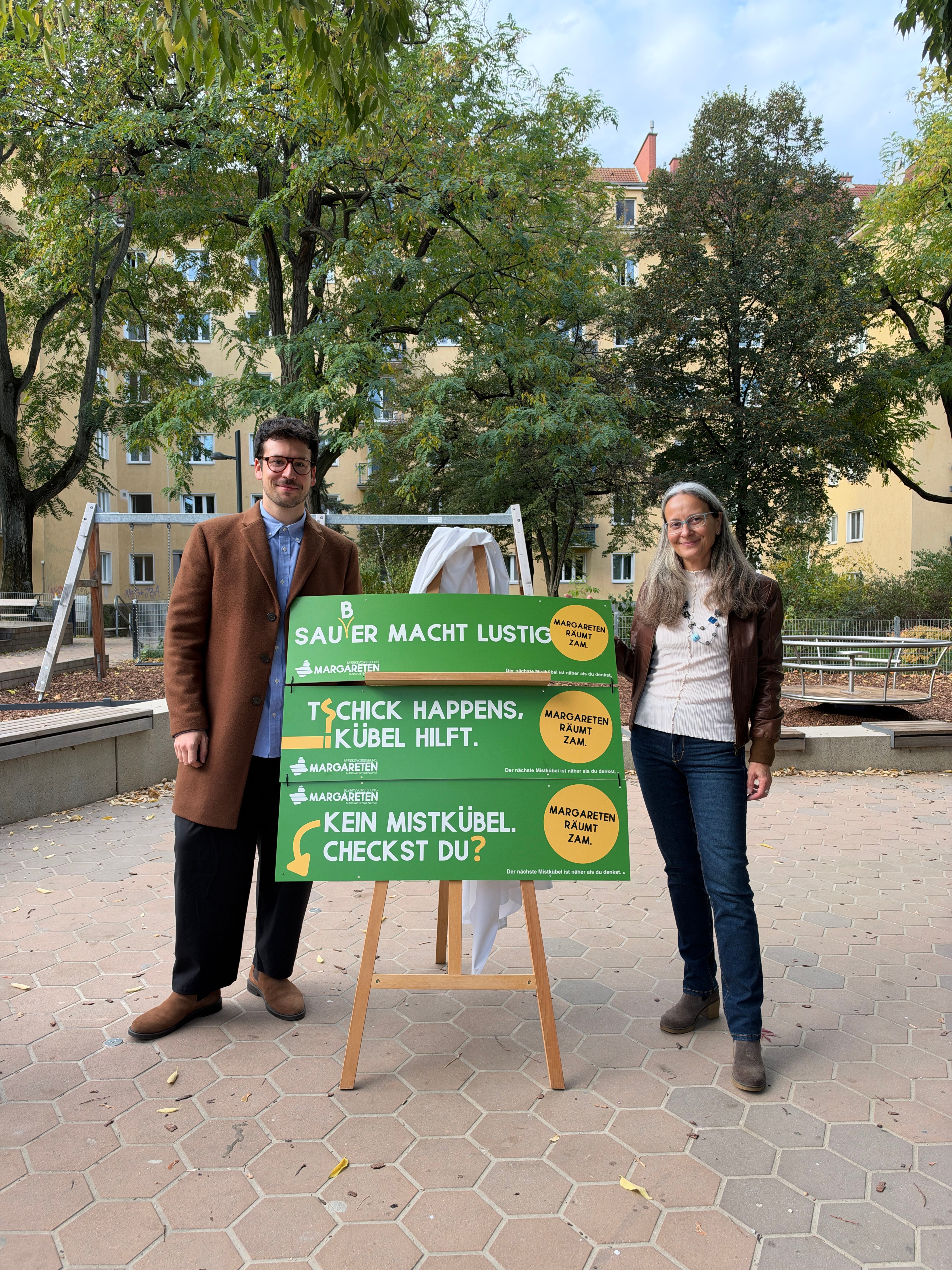 Zwei Personen stehen neben einer Tafel mit Slogans zur Anti-Müll-Kampagne "Margareten räumt zam".