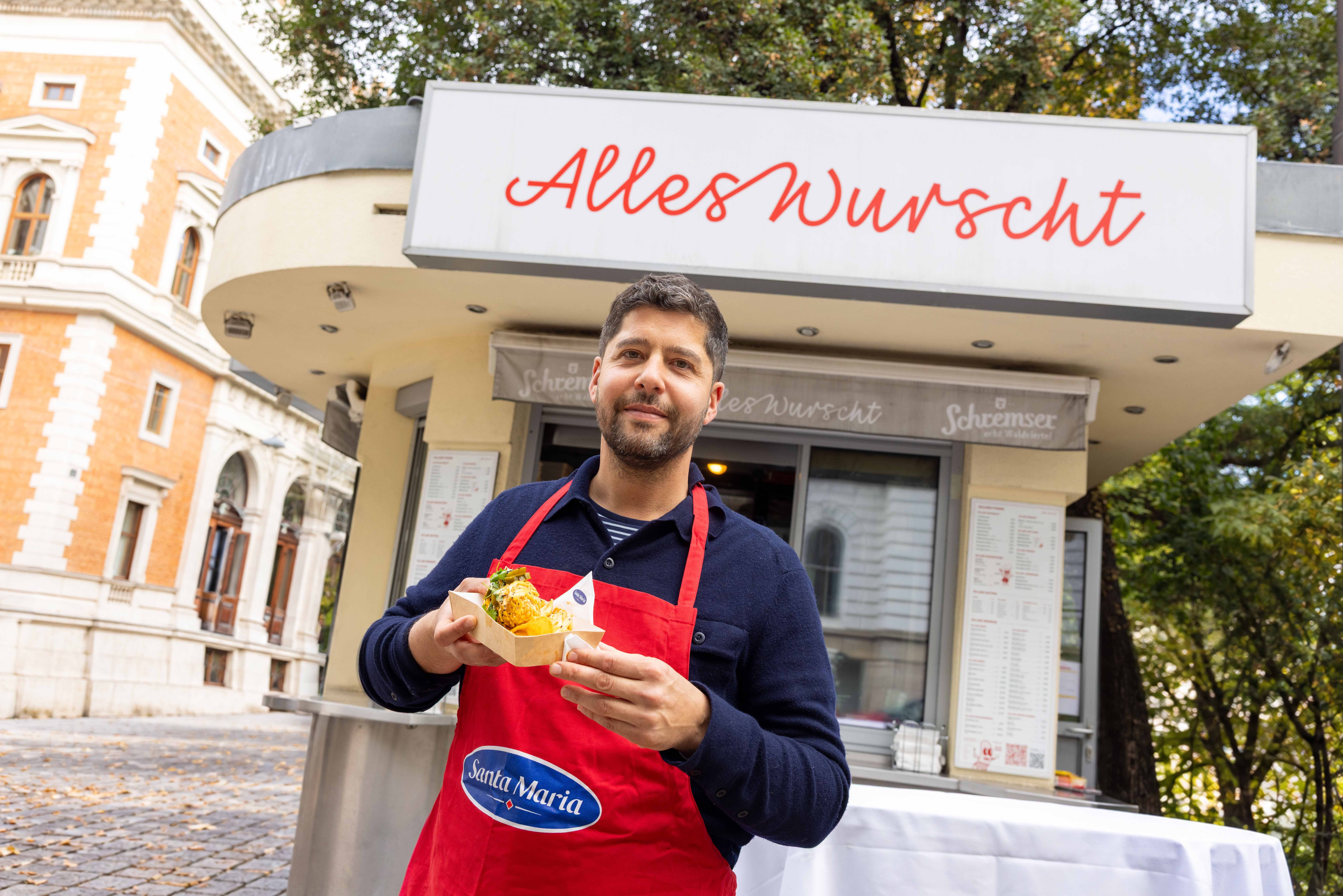 Mann mit roter Schürze hält einen Wiener Nacho Dog vor einem Imbissstand namens "Alles Wurscht".