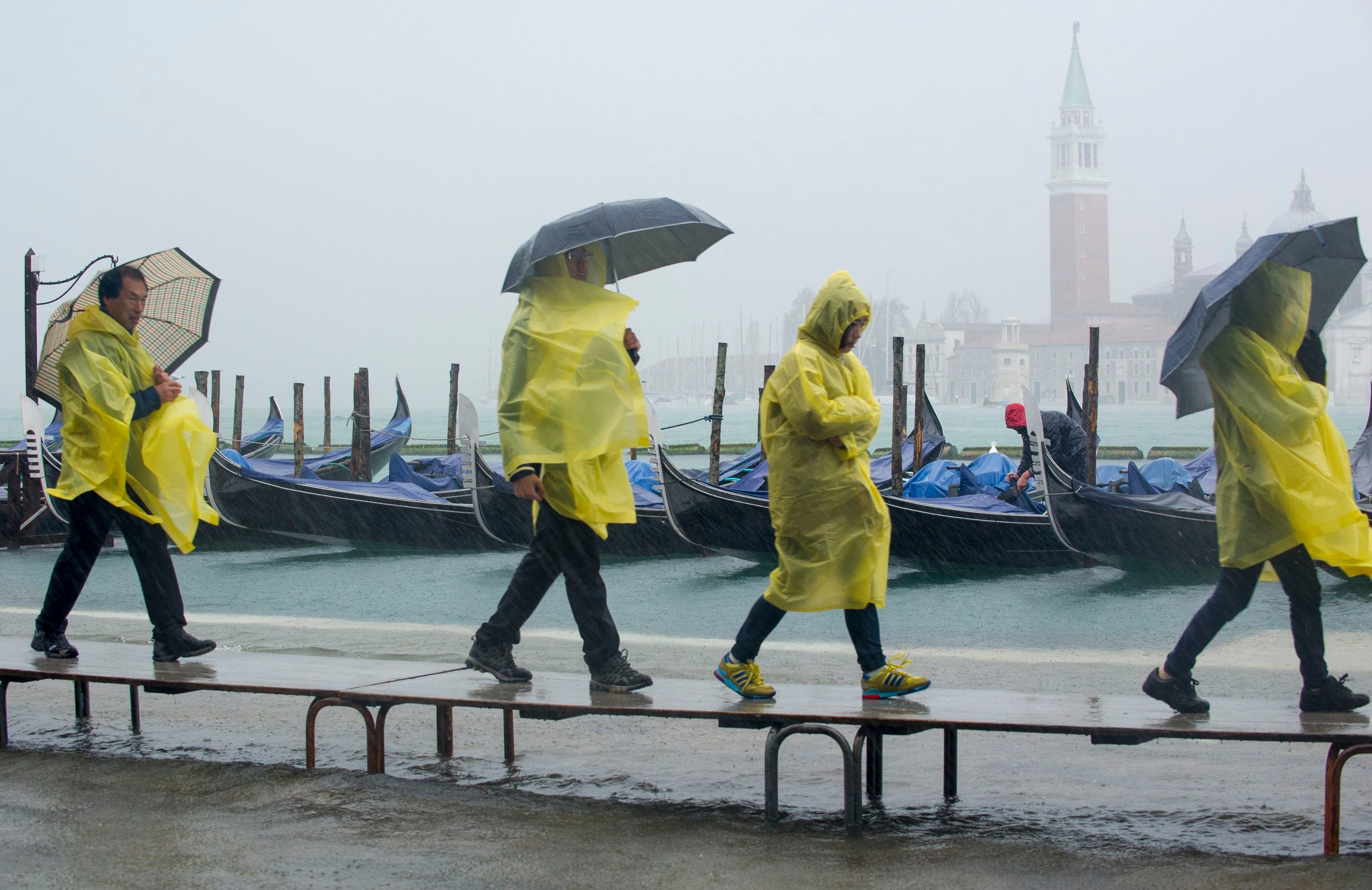 Touristen in gelben Regenponchos gehen bei Regen mit Regenschirmen an überfluteten Gondeln vorbei.