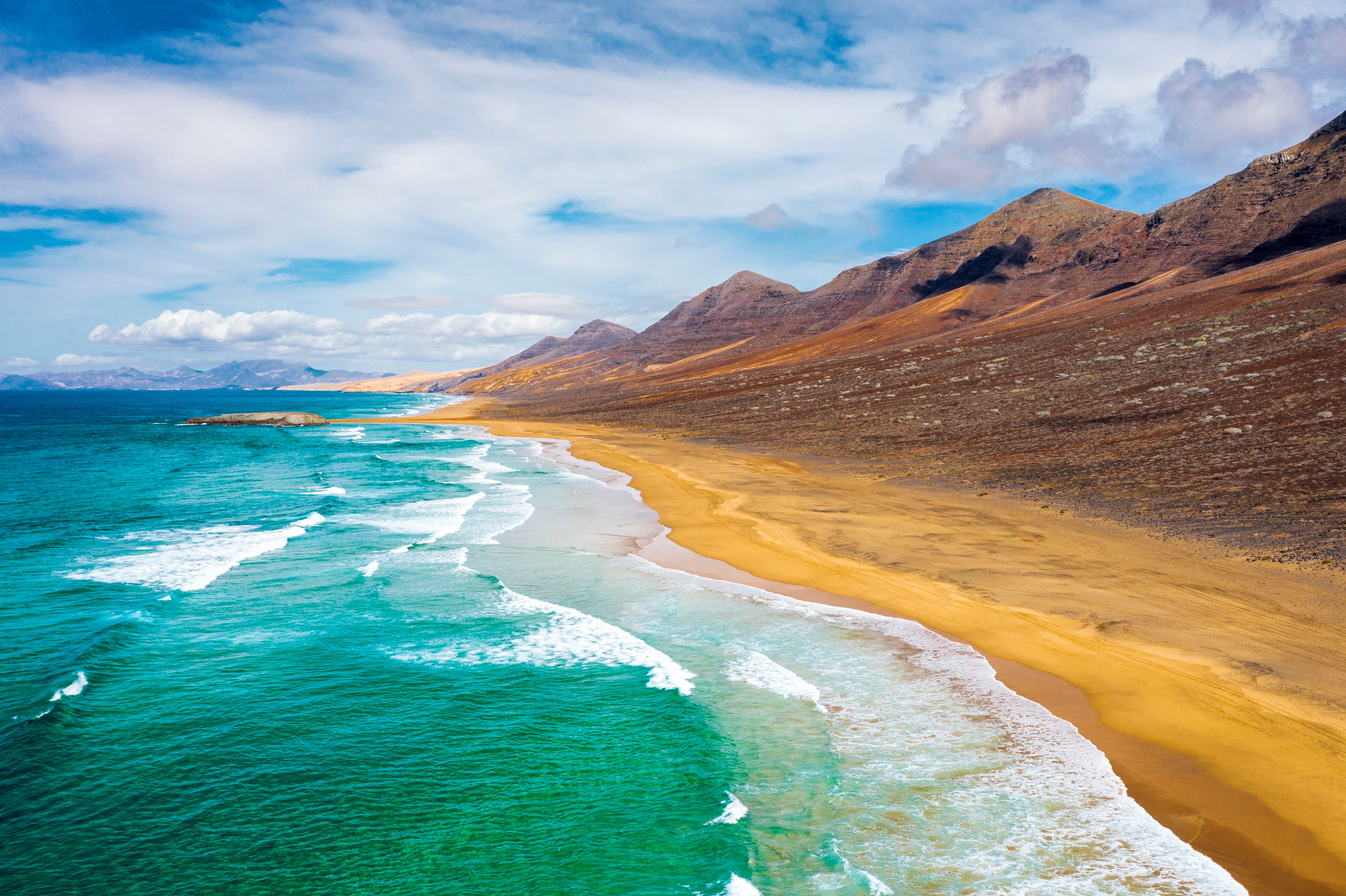 Wellen treffen auf den goldenen Sandstrand von Cofete mit Bergen im Hintergrund.