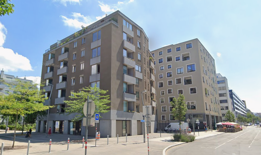 Mehrstöckiges Wohnhaus an einer Straßenecke mit Bäumen und Gehwegen bei Tag.