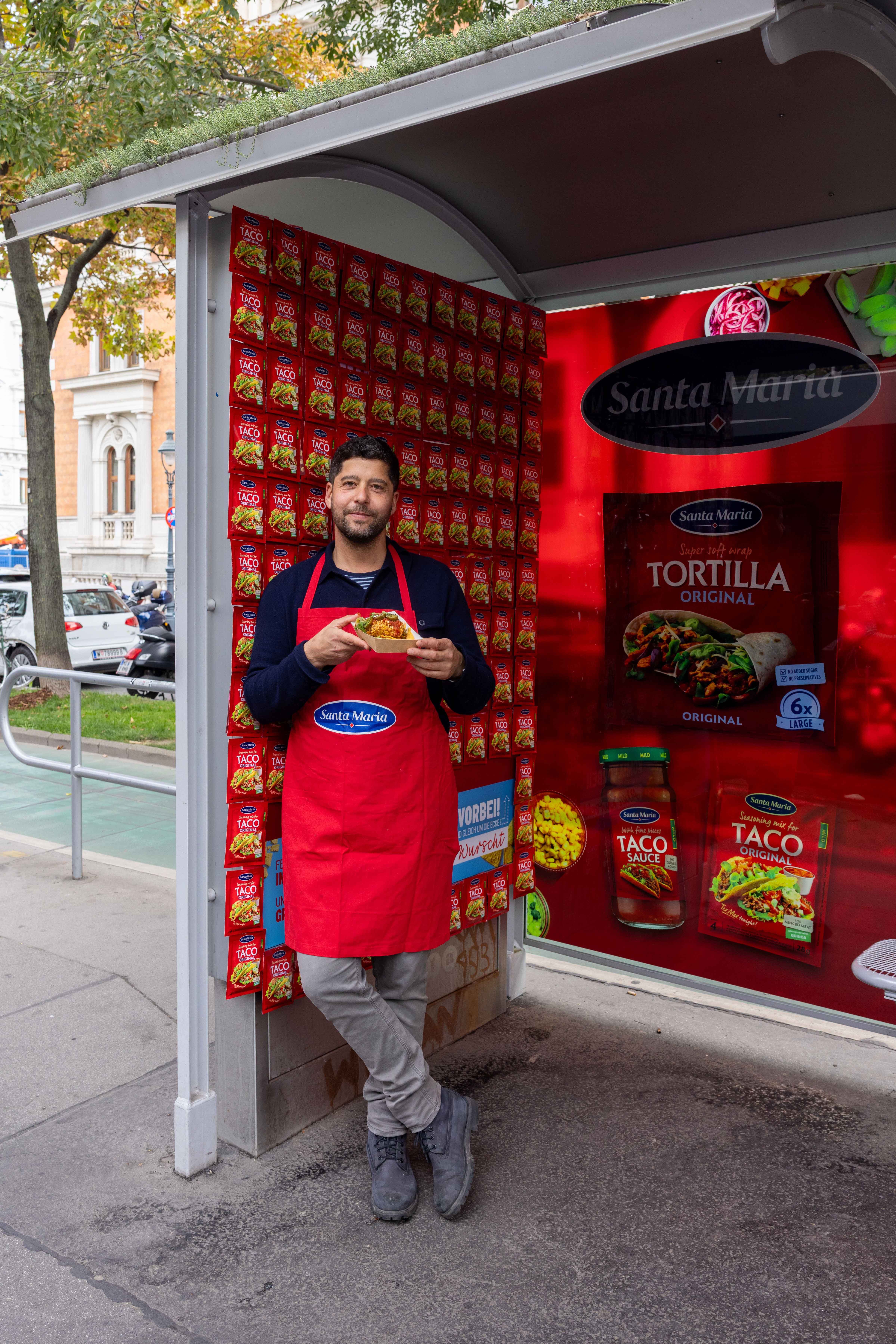 Ein Mann mit roter Schürze hält einen Nacho Dog an einer bunten Santa Maria Werbewand.