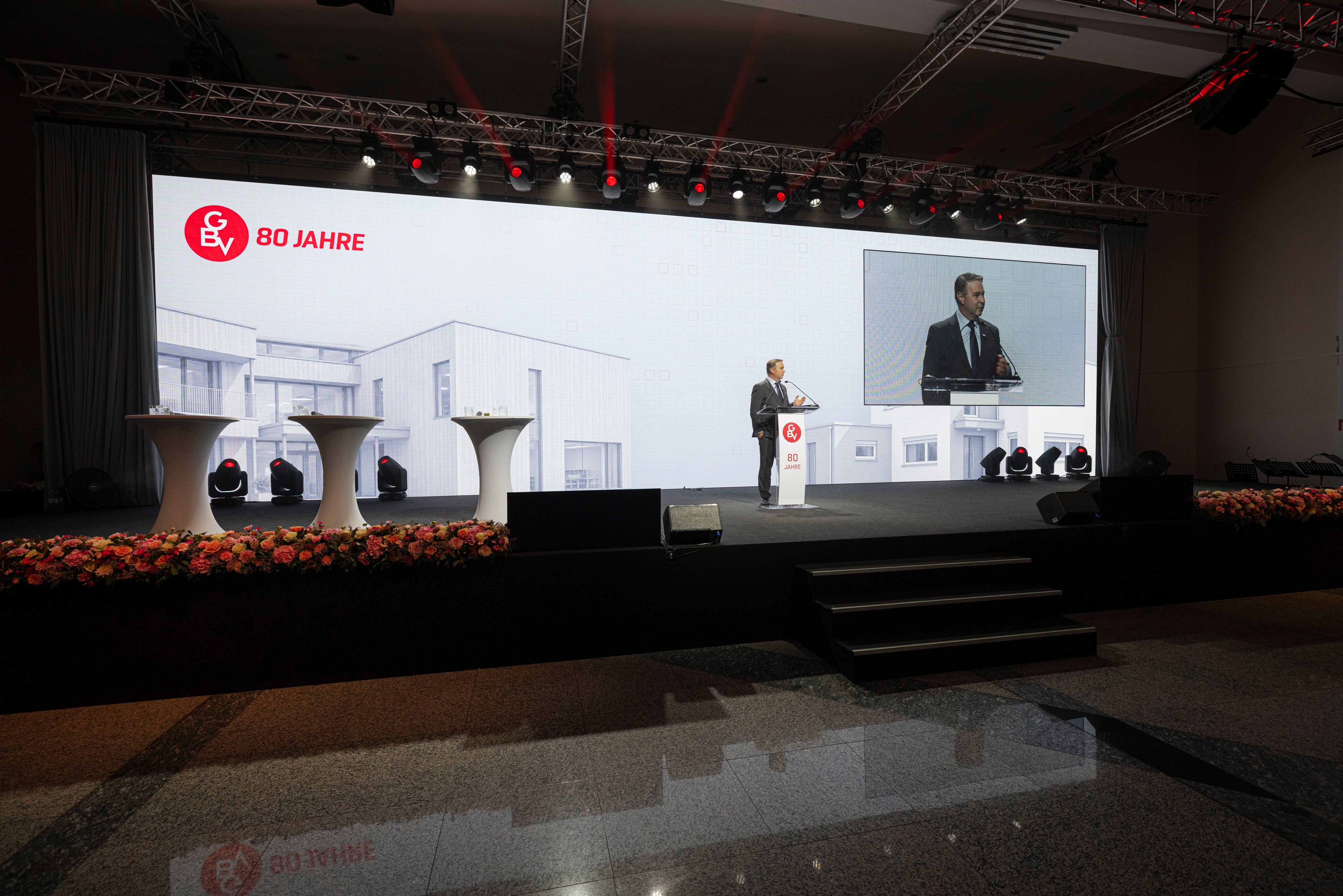 Bühne mit Rednerpult, Mann hält Rede, im Hintergrund große Leinwand mit „80 Jahre“-Logo.