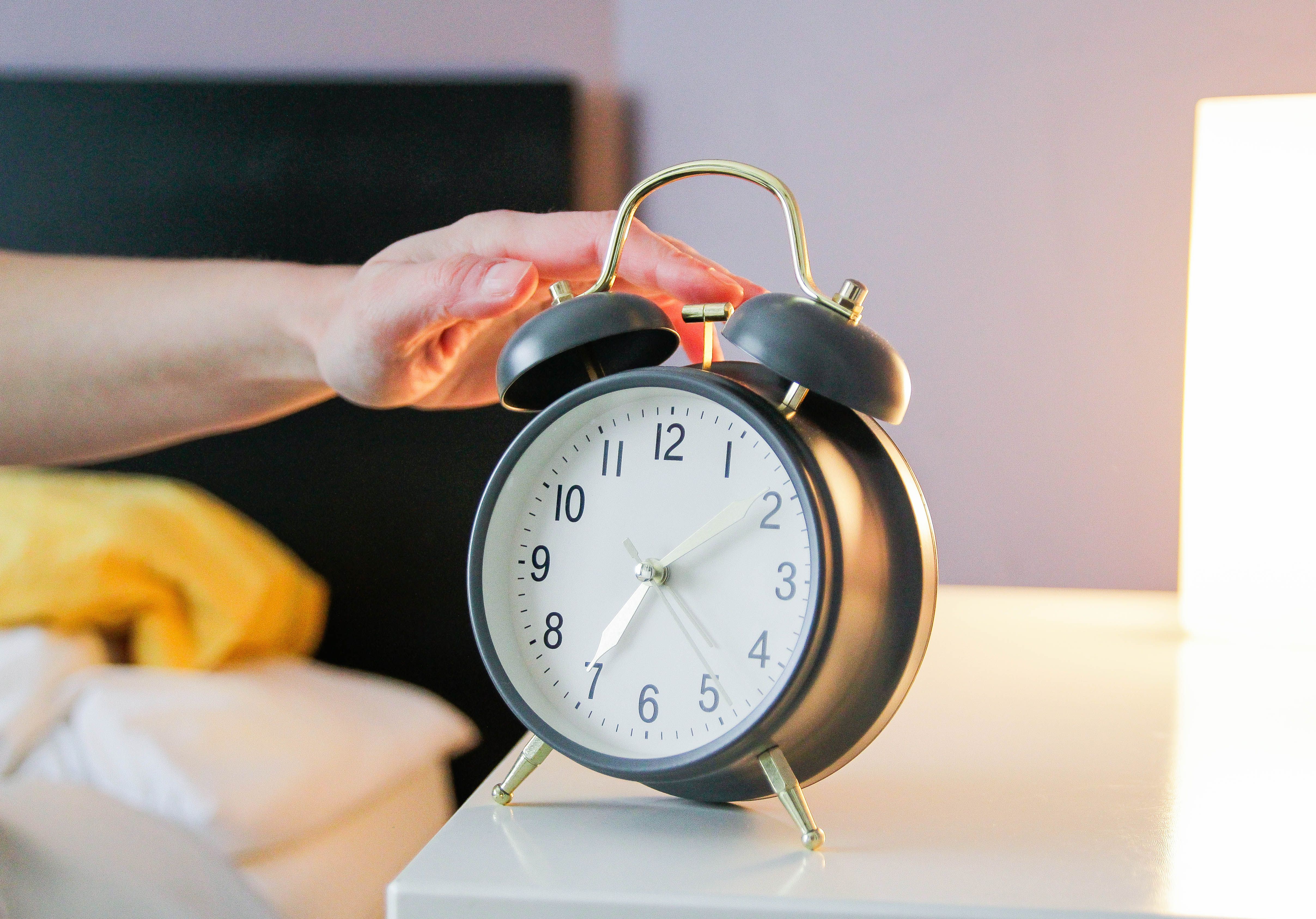 Hand schaltet einen schwarzen Wecker auf einem Nachttisch neben einem Bett aus.