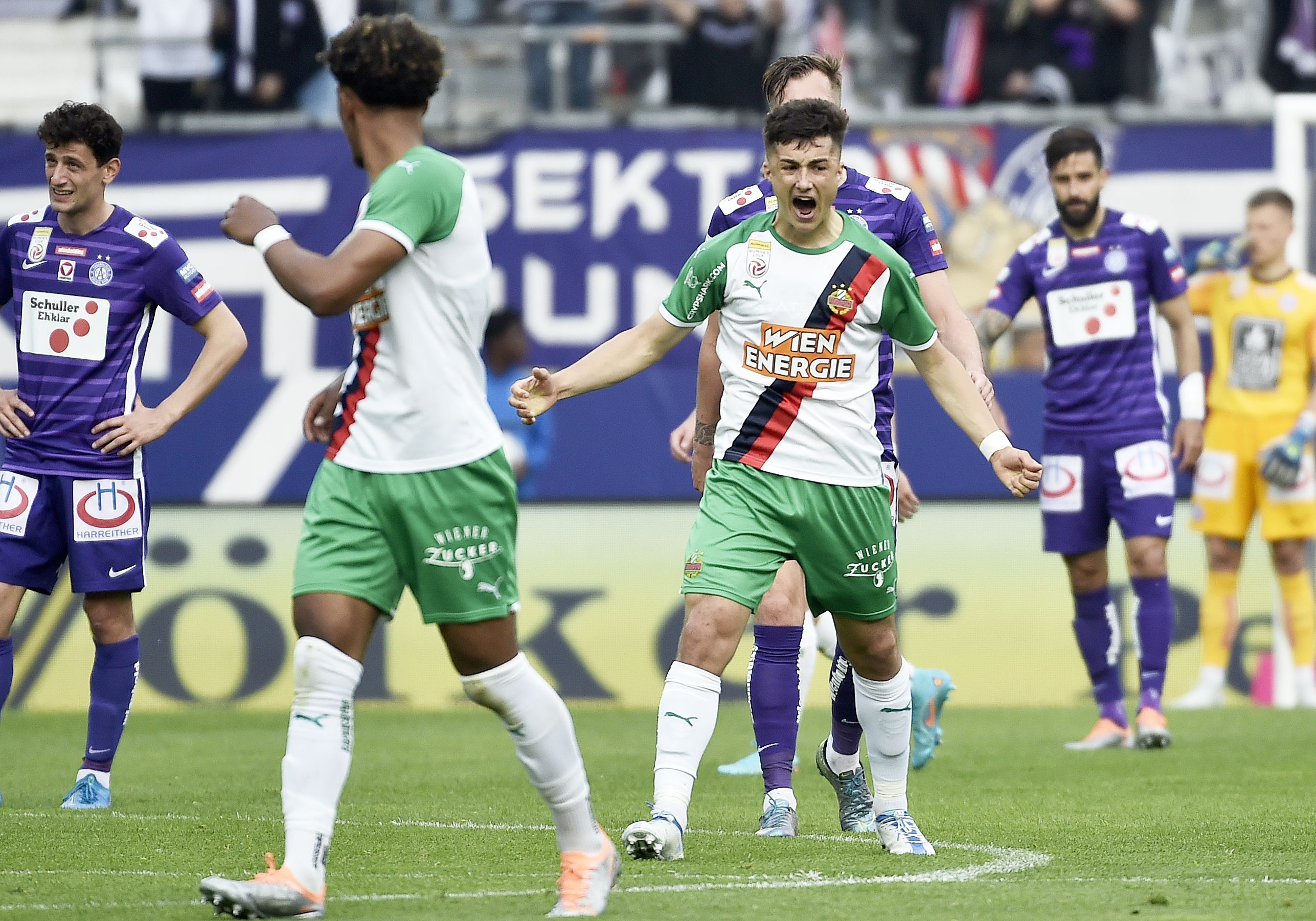 Bernhard Zimmermann im grün-weißen Rapid-Trikot schreit mit geballten Fäusten vor Enttäuschung auf dem Rasen, umgeben von enttäuschten Mitspielern und gegnerischen Spielern der Austria.