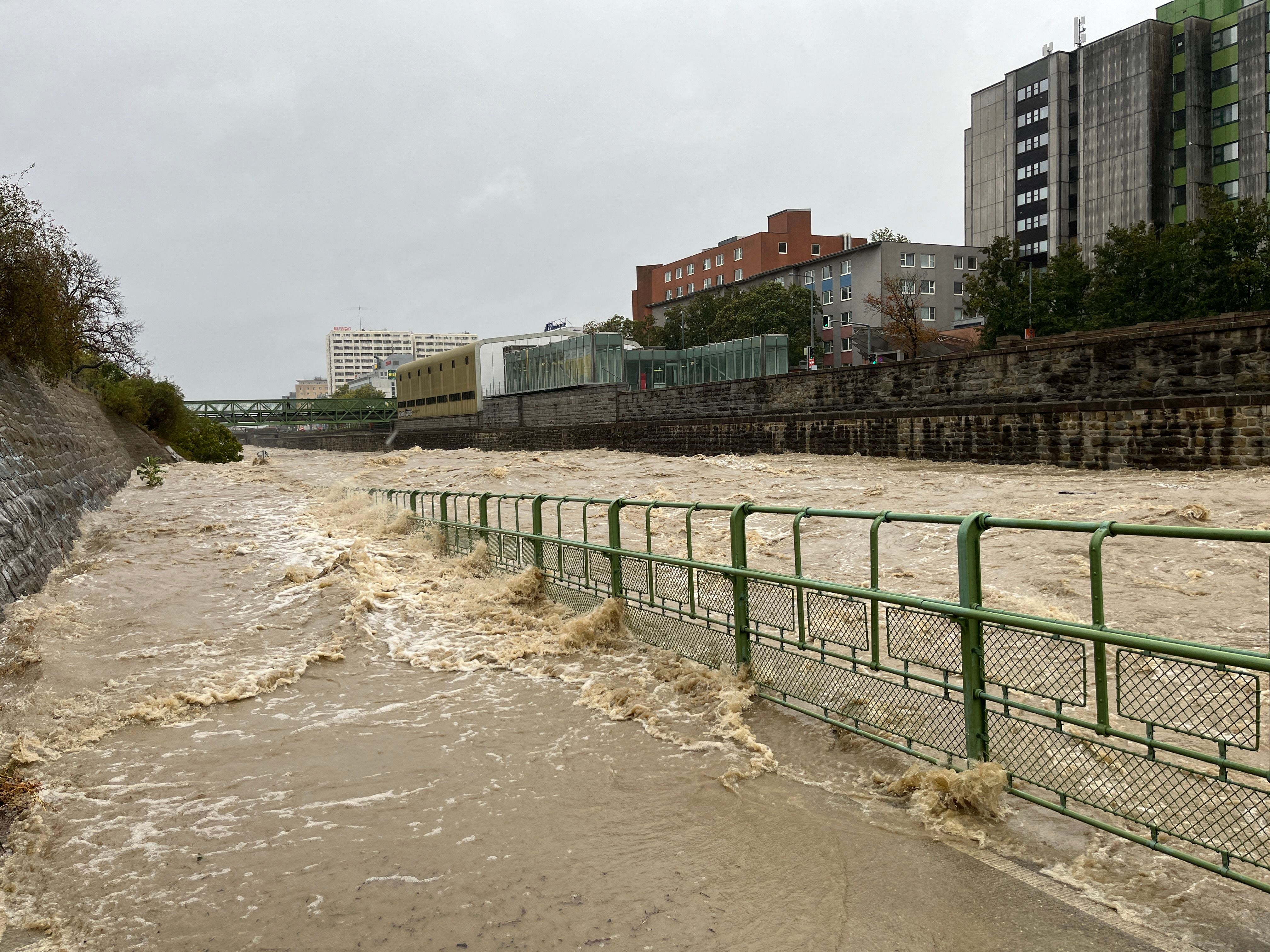 Überfluteter Fluss mit Hochwasserschutz-Geländer in einer Stadt bei starkem Regen.