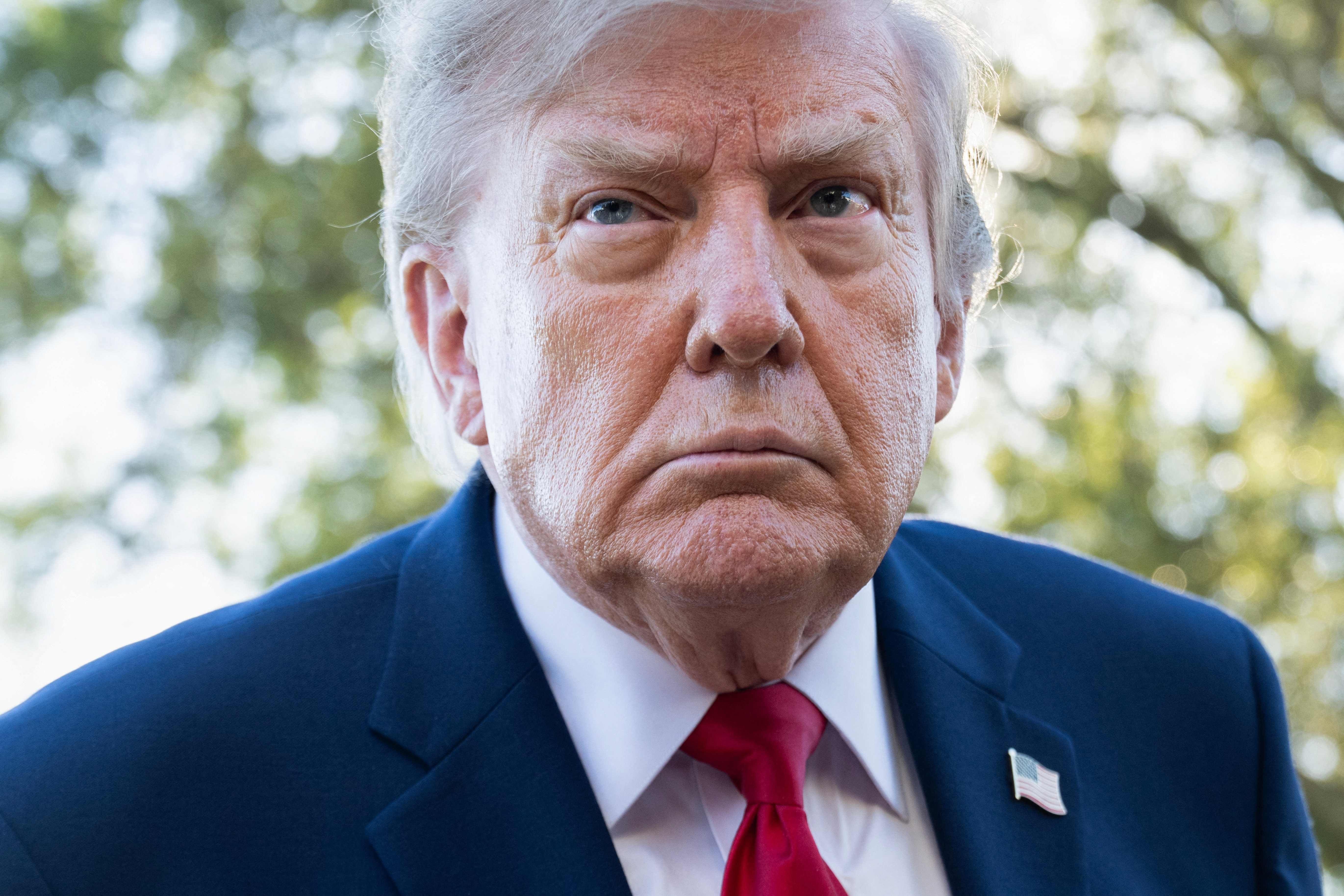 ALT-Text Nahaufnahme von US-Präsident Donald Trump in blauem Anzug und roter Krawatte mit ernstem Gesichtsausdruck vor grünem Hintergrund im Freien.