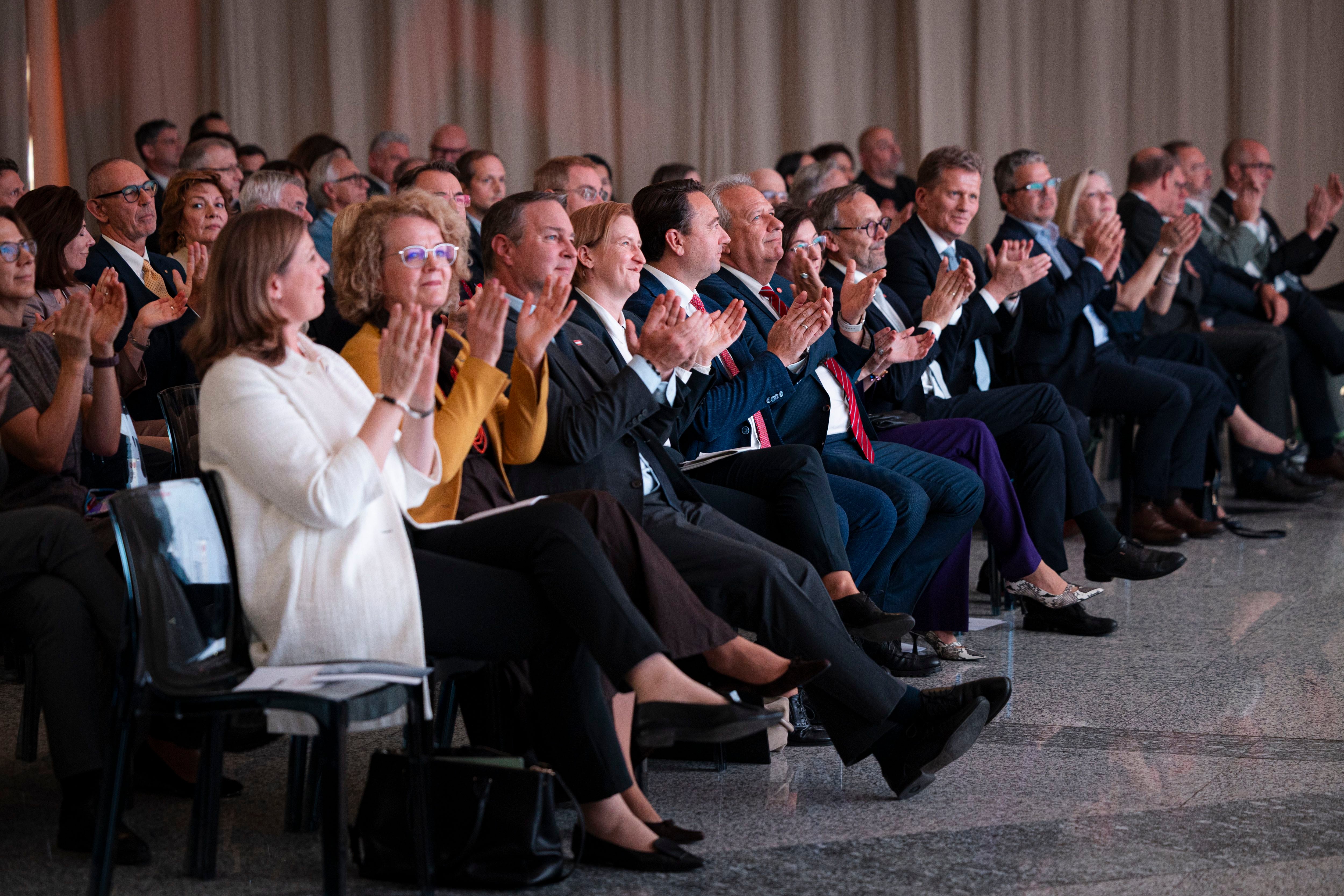 Menschen sitzen in Reihen und applaudieren auf einer Veranstaltung in einem Saal.