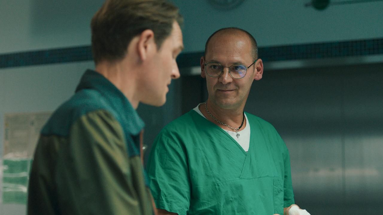 Zwei Männer stehen in einem Raum, einer trägt grüne medizinische Kleidung und eine Brille.