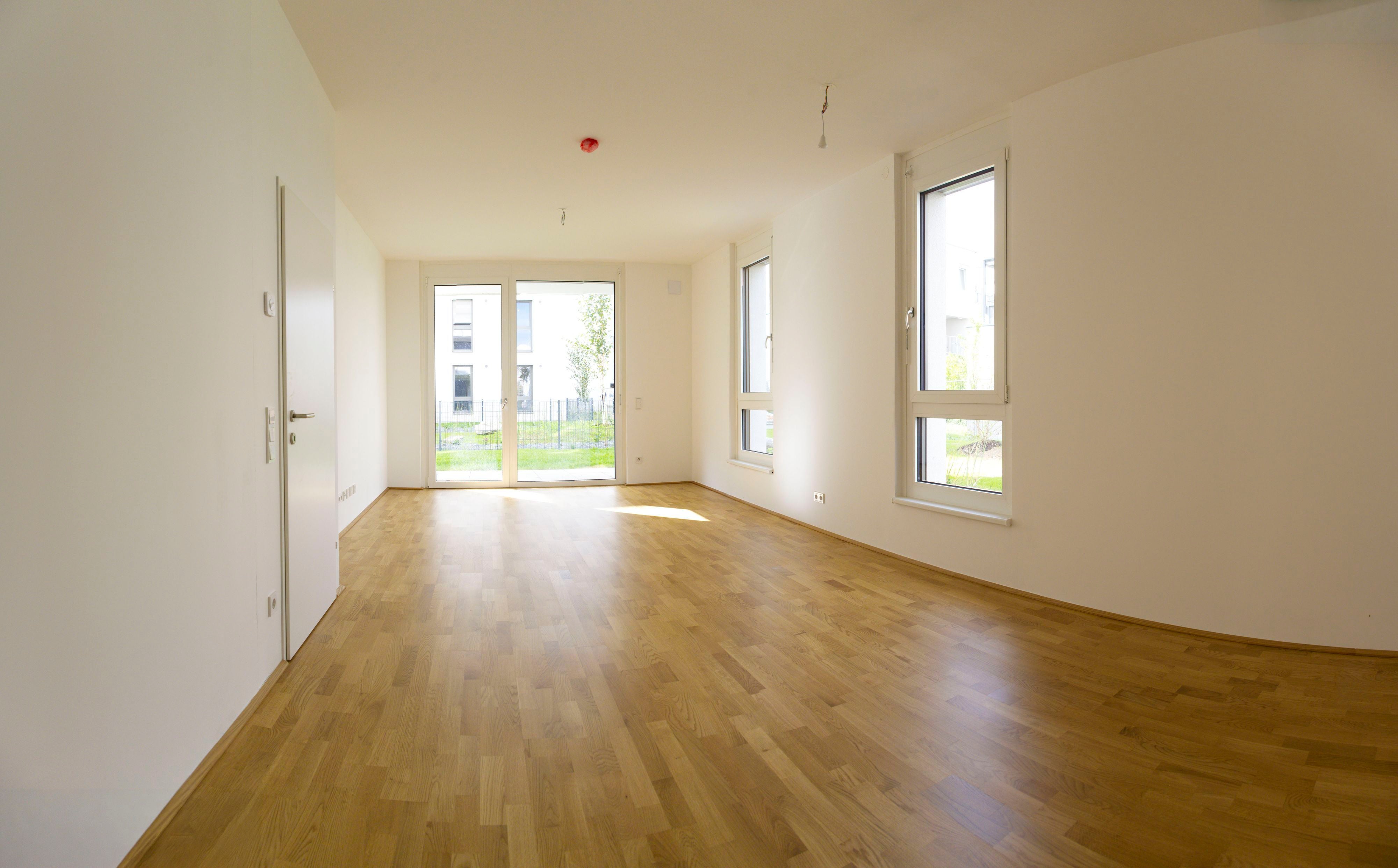 Heller, leerer Raum mit Parkettboden, weißen Wänden und großen Fenstern zur Terrasse.