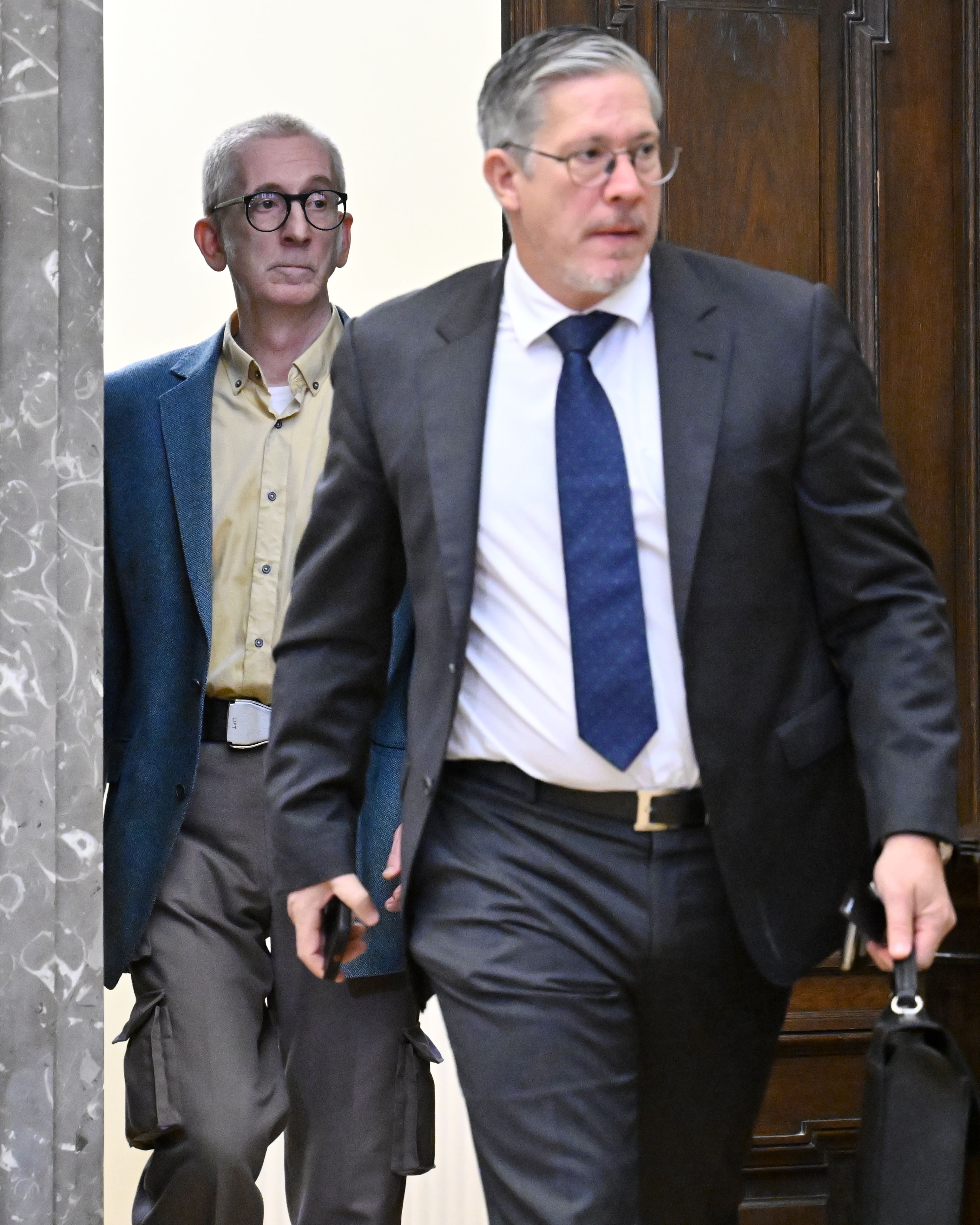 Zwei Männer gehen in einem Gerichtsgebäude, einer trägt Anzug, der andere Sakko und Brille.