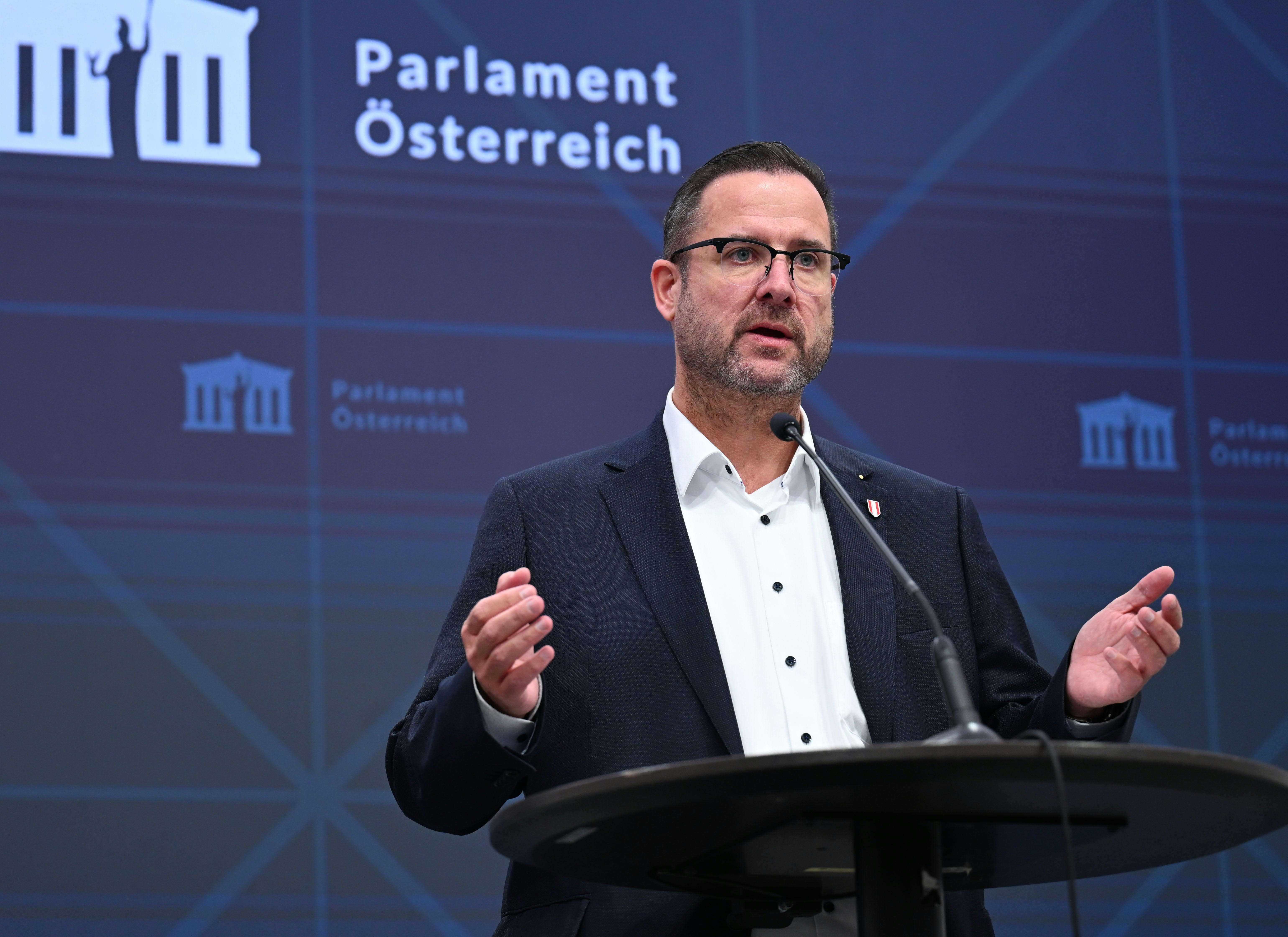 Ein Mann spricht am Rednerpult im Parlament Österreich, Hintergrund mit blauem Logo.