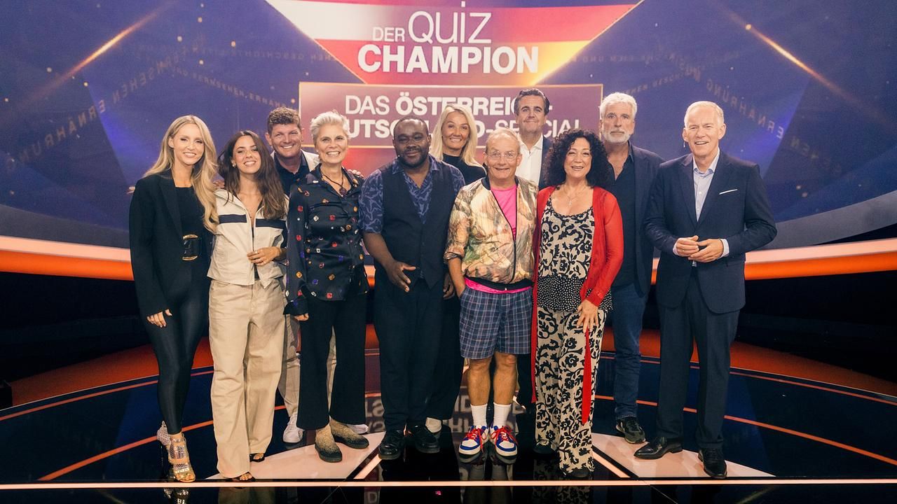 Gruppenfoto von elf Menschen auf einer Quizshow-Bühne mit dem Schriftzug „Der Quiz Champion“.