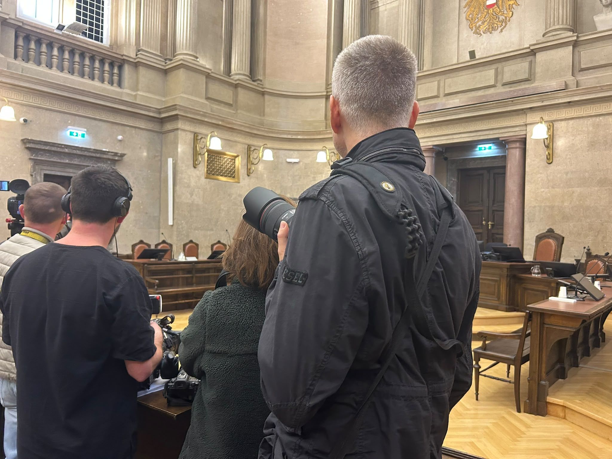 Mehrere Personen mit Kameras und Mikrofonen stehen in einem großen Gerichtssaal.