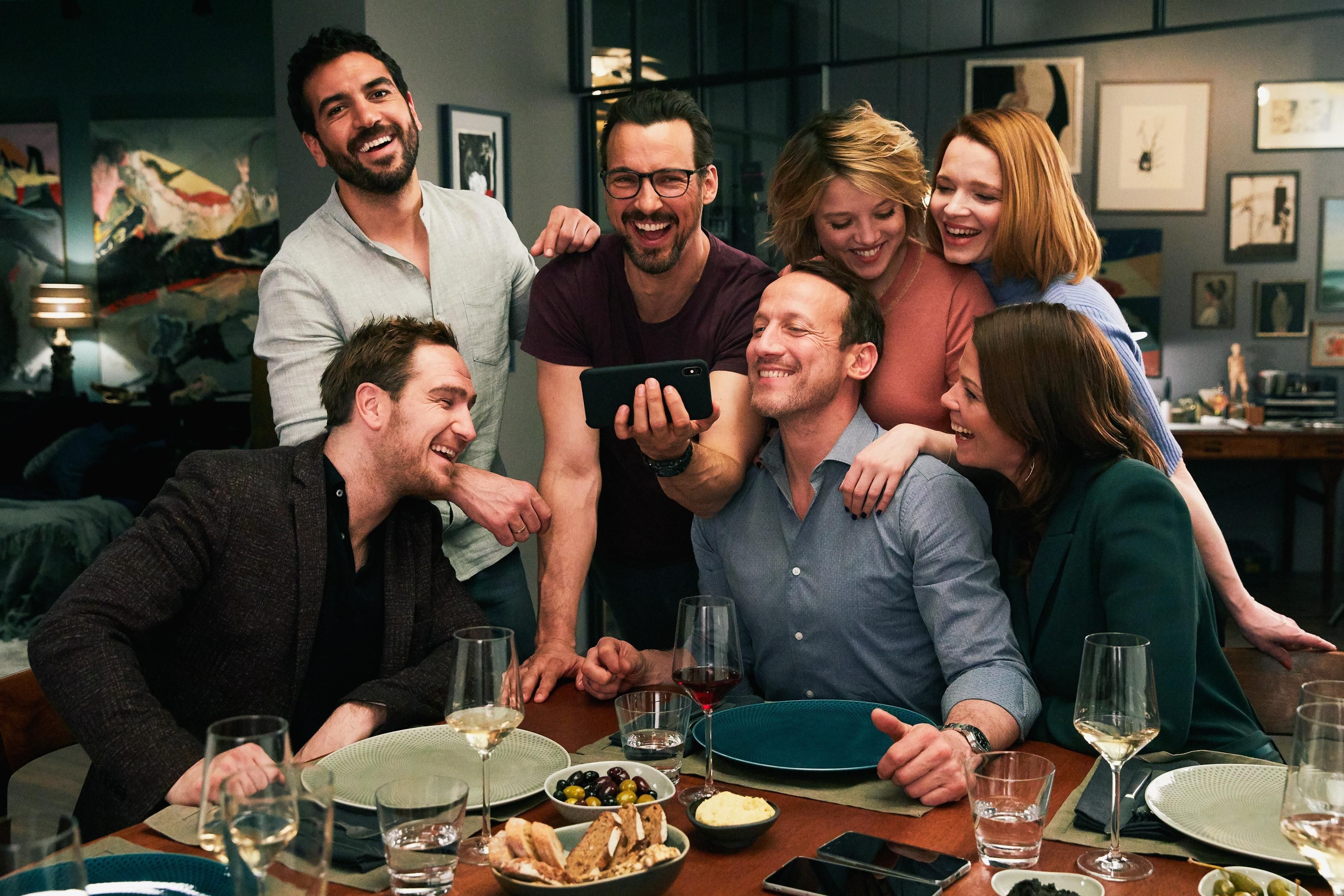 Sieben Freunde sitzen lächelnd um einen Tisch und schauen gemeinsam auf ein Smartphone.