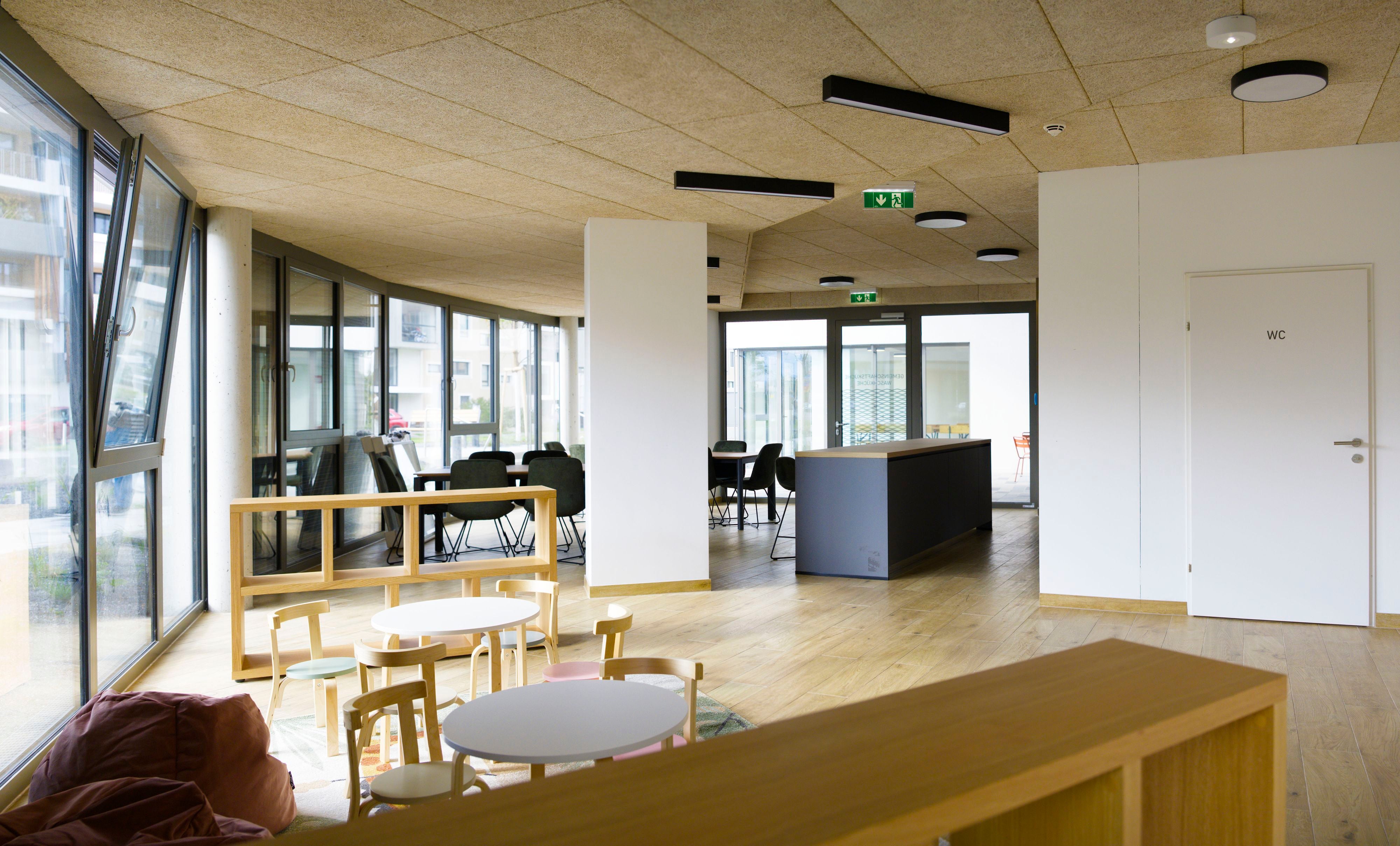 Heller Gemeinschaftsraum mit Kindertischen, Sitzsäcken, großen Fenstern und einer Theke.