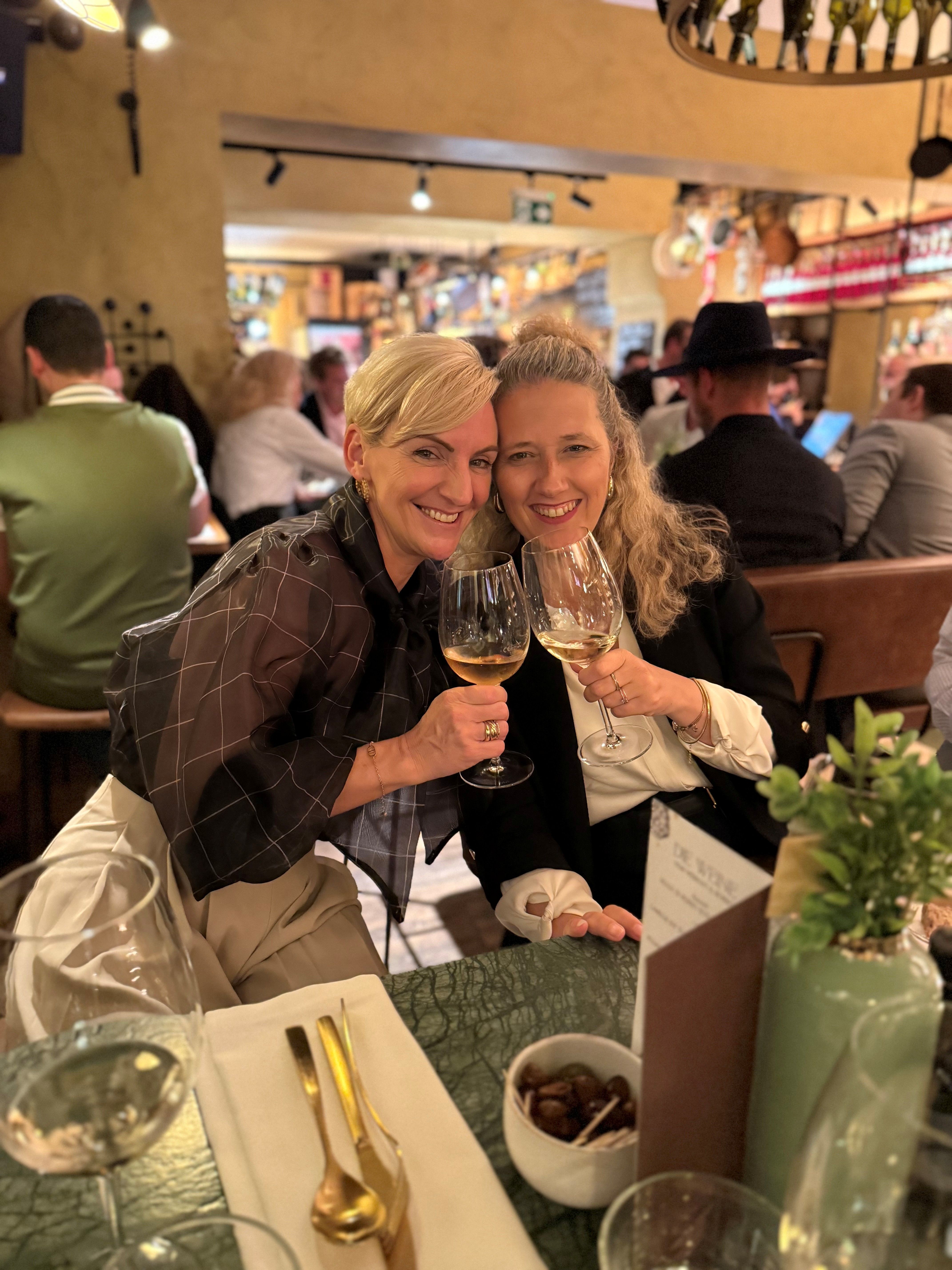 Zwei Frauen stoßen in einem Restaurant mit Weingläsern an und lächeln in die Kamera.
