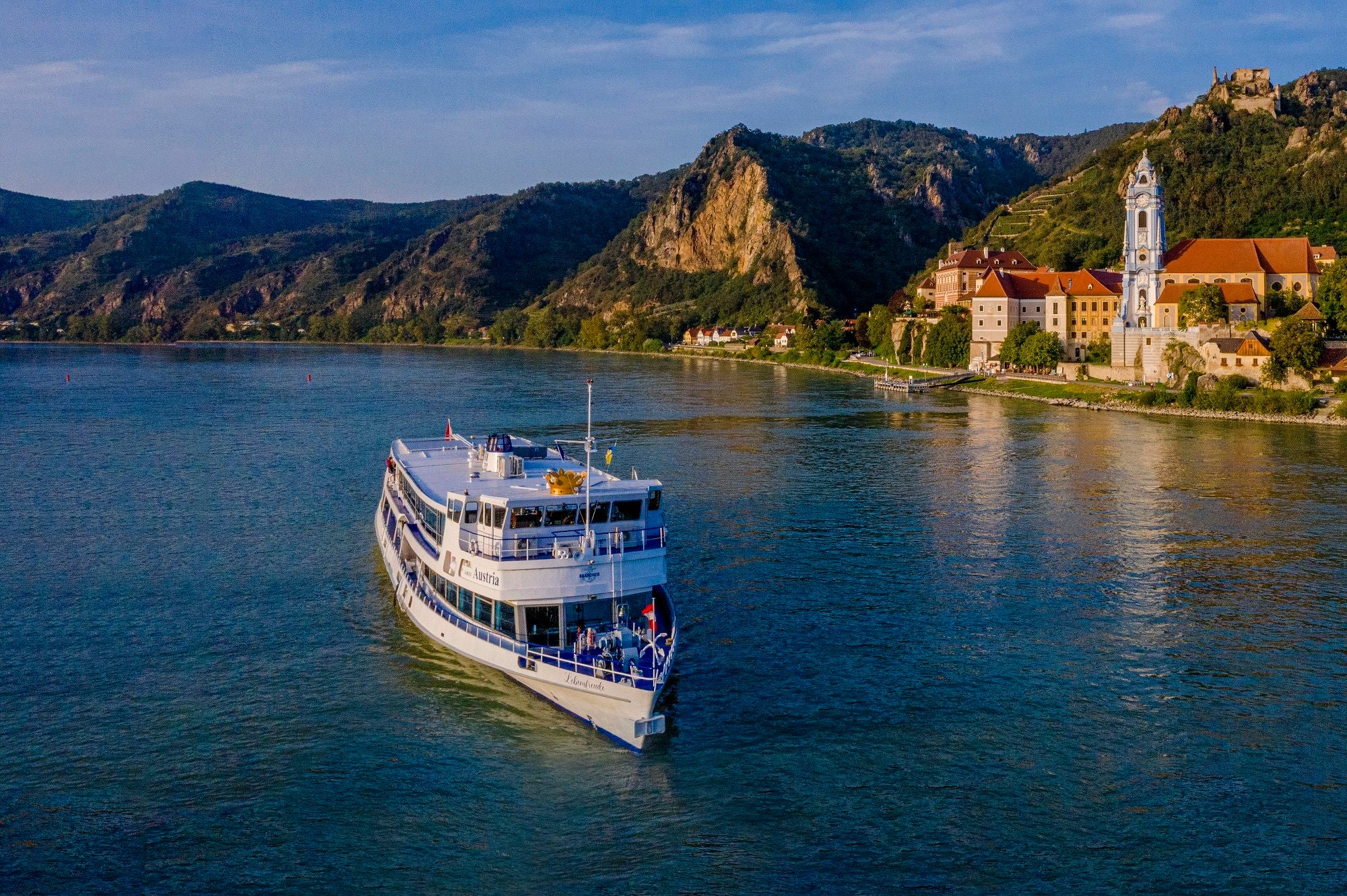 Personenschiff MS Austria fährt auf der Donau vor Dürnstein, im Hintergrund Berge und Kirche.