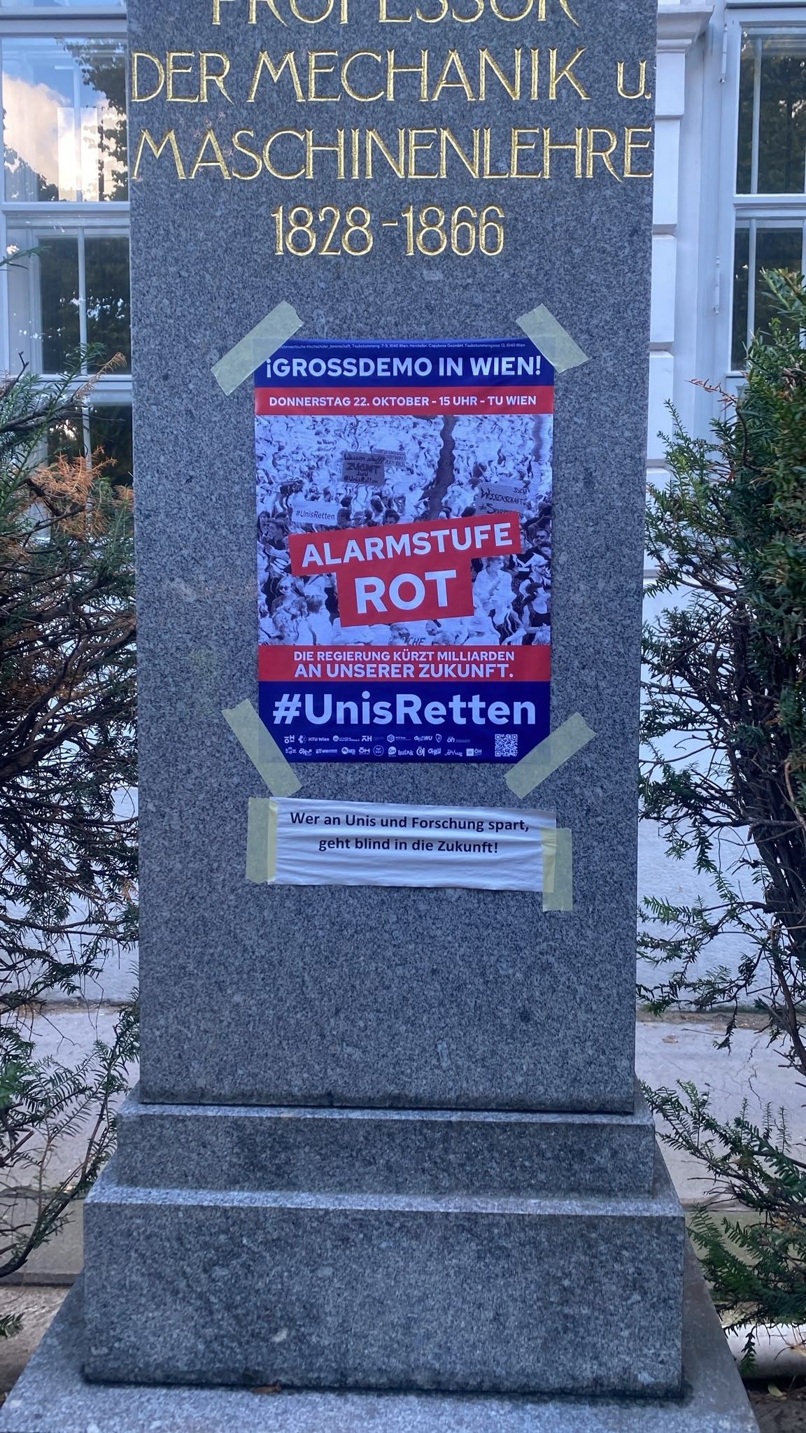 Plakat mit "Alarmstufe Rot" und #UnisRetten auf einem Denkmal, Demoankündigung in Wien.