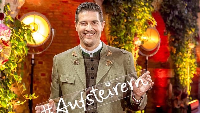 Mann in Tracht hält Schild mit dem Schriftzug "#Aufsteiern" vor bunter, festlicher Kulisse.