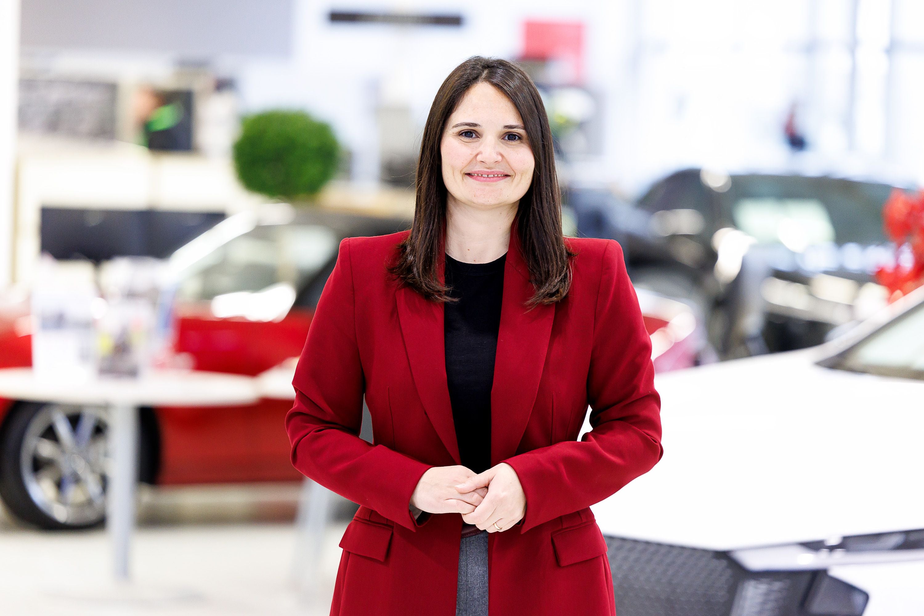 Frau mit rotem Blazer steht lächelnd in einem Autohaus zwischen ausgestellten Autos.