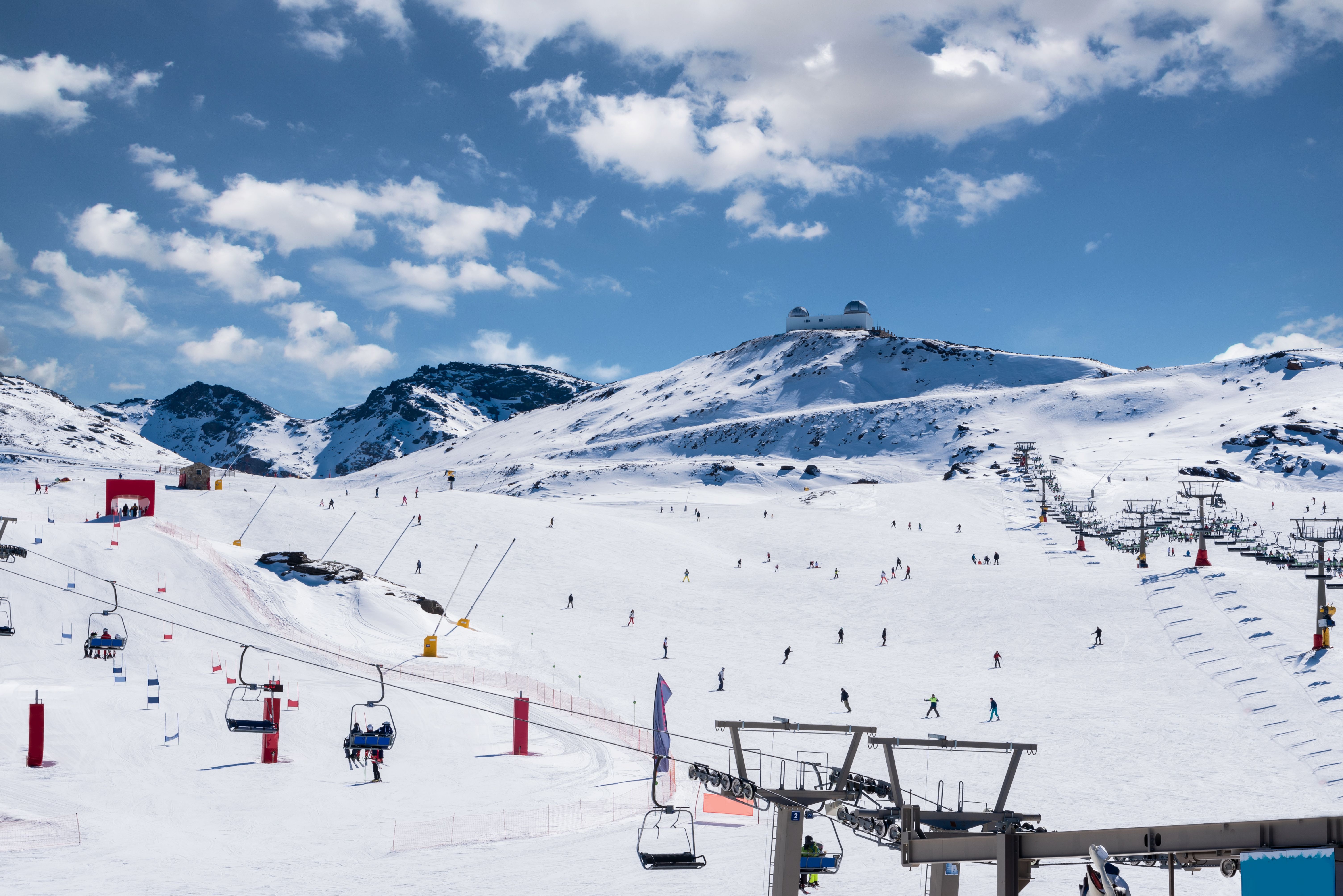 Skigebiet Sierra Nevada mit Sesselliften, Skifahrern und Observatorium im Hintergrund bei Sonnenschein.