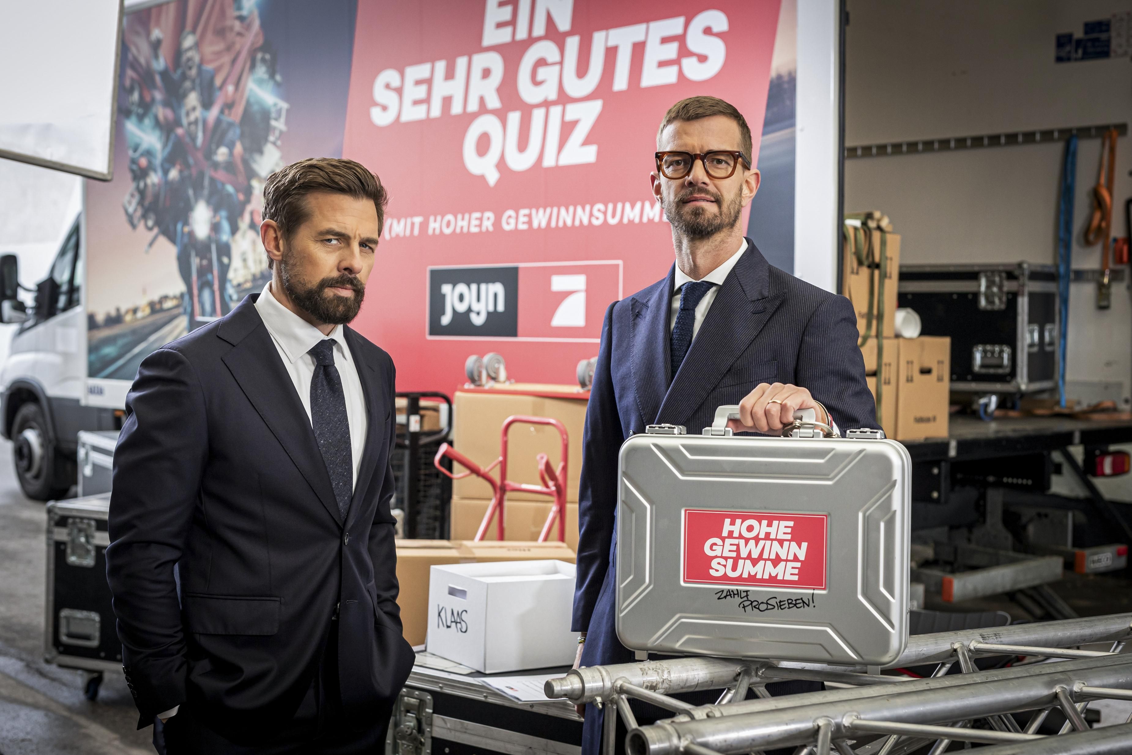 Zwei Männer in Anzügen stehen vor einem Koffer mit der Aufschrift "Hohe Gewinnsumme".