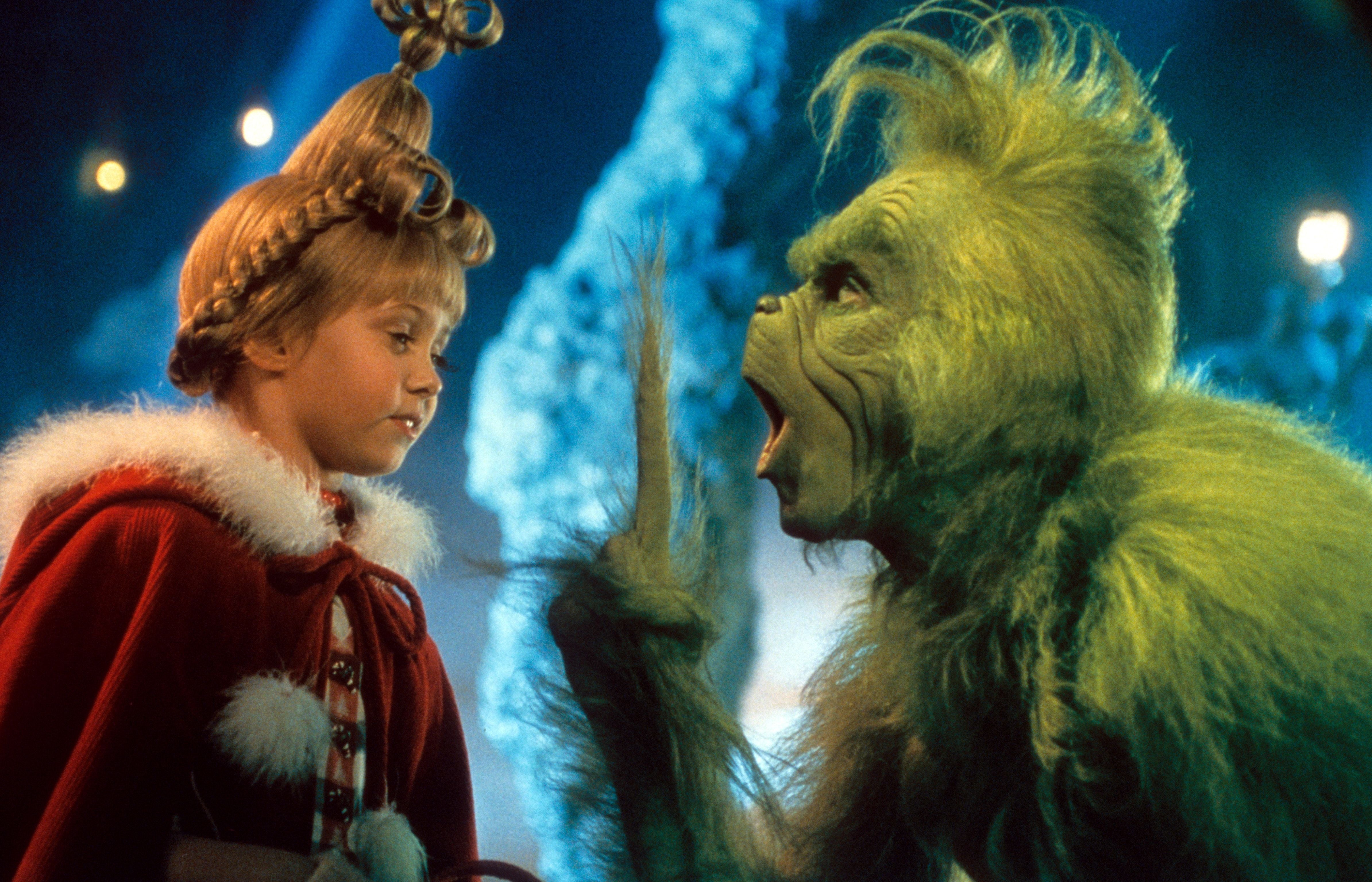 Ein Mädchen im roten Umhang schaut den grünen Grinch an, der den Mund weit geöffnet hat.