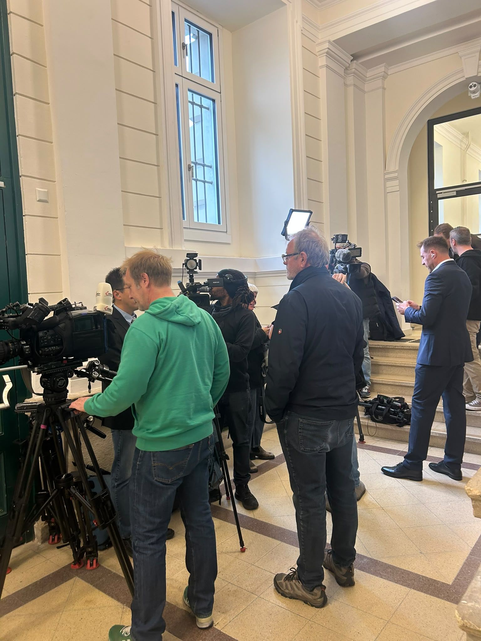 Mehrere Kamerateams und Reporter filmen und arbeiten in einem hellen, klassisch gestalteten Flur.