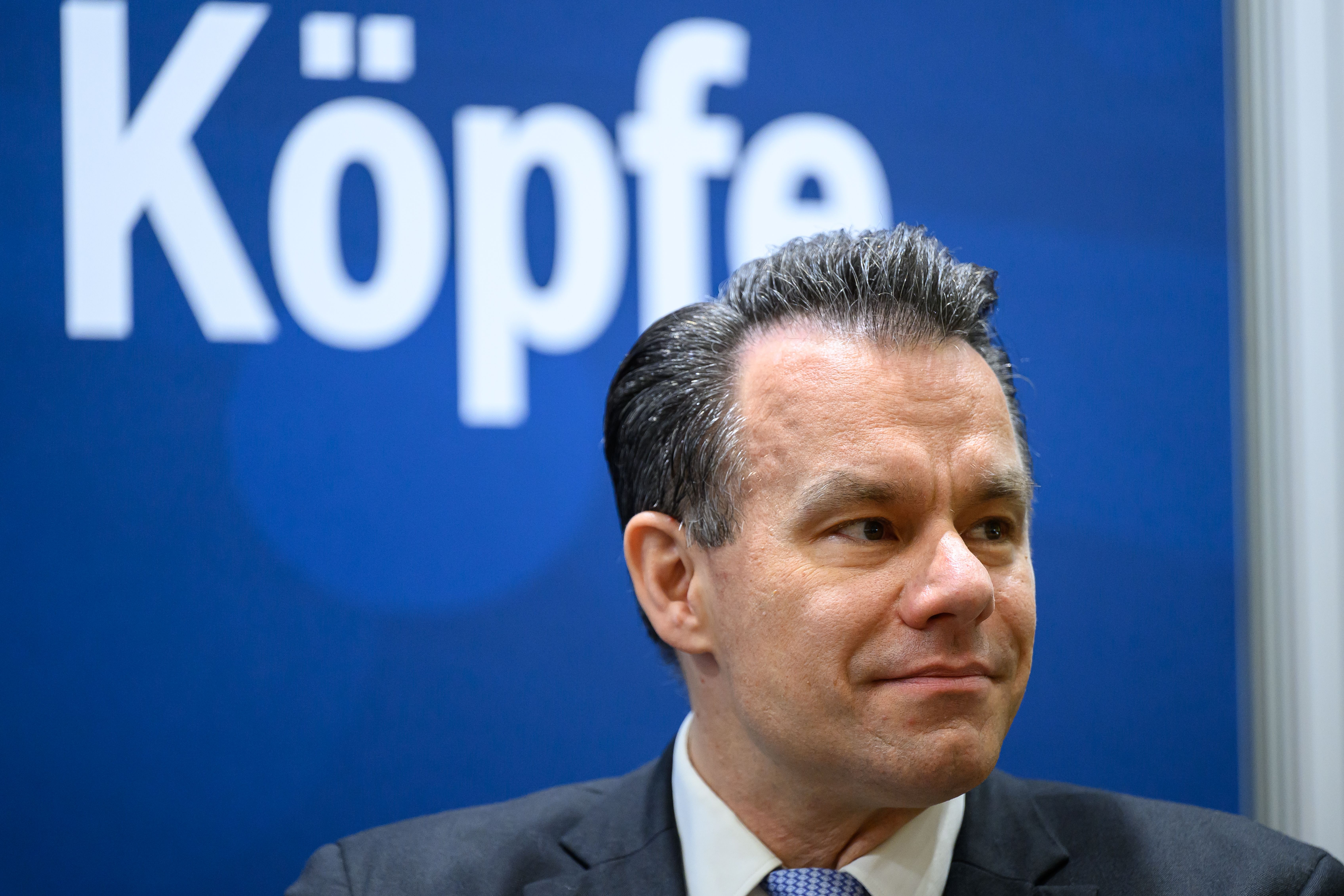 Ein Mann im Anzug sitzt vor einer blauen Wand mit dem Wort "Köpfe" im Hintergrund.