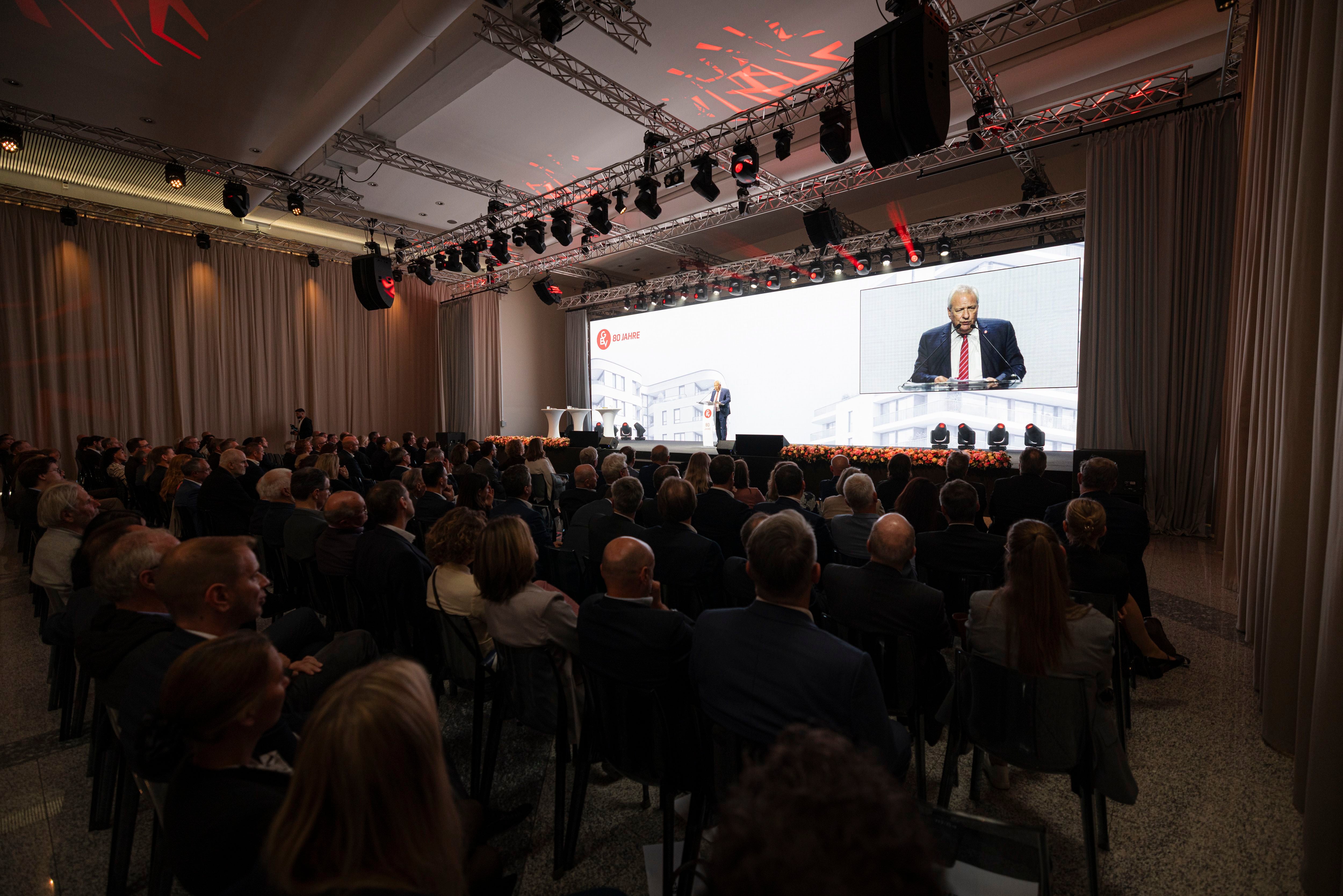 Ein Mann hält eine Rede auf einer Bühne vor großem Publikum, mit Projektion im Hintergrund.