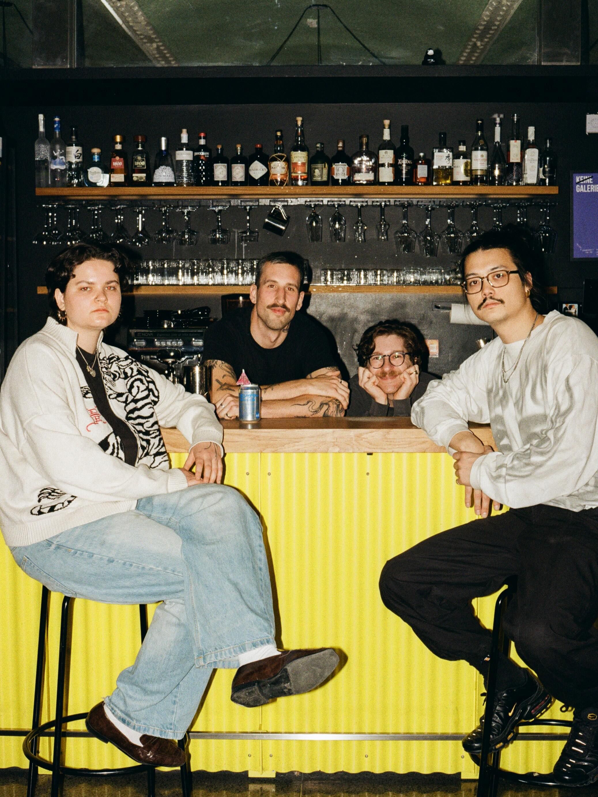Vier Personen sitzen und stehen an einer Bar mit Flaschen und Gläsern im Hintergrund.