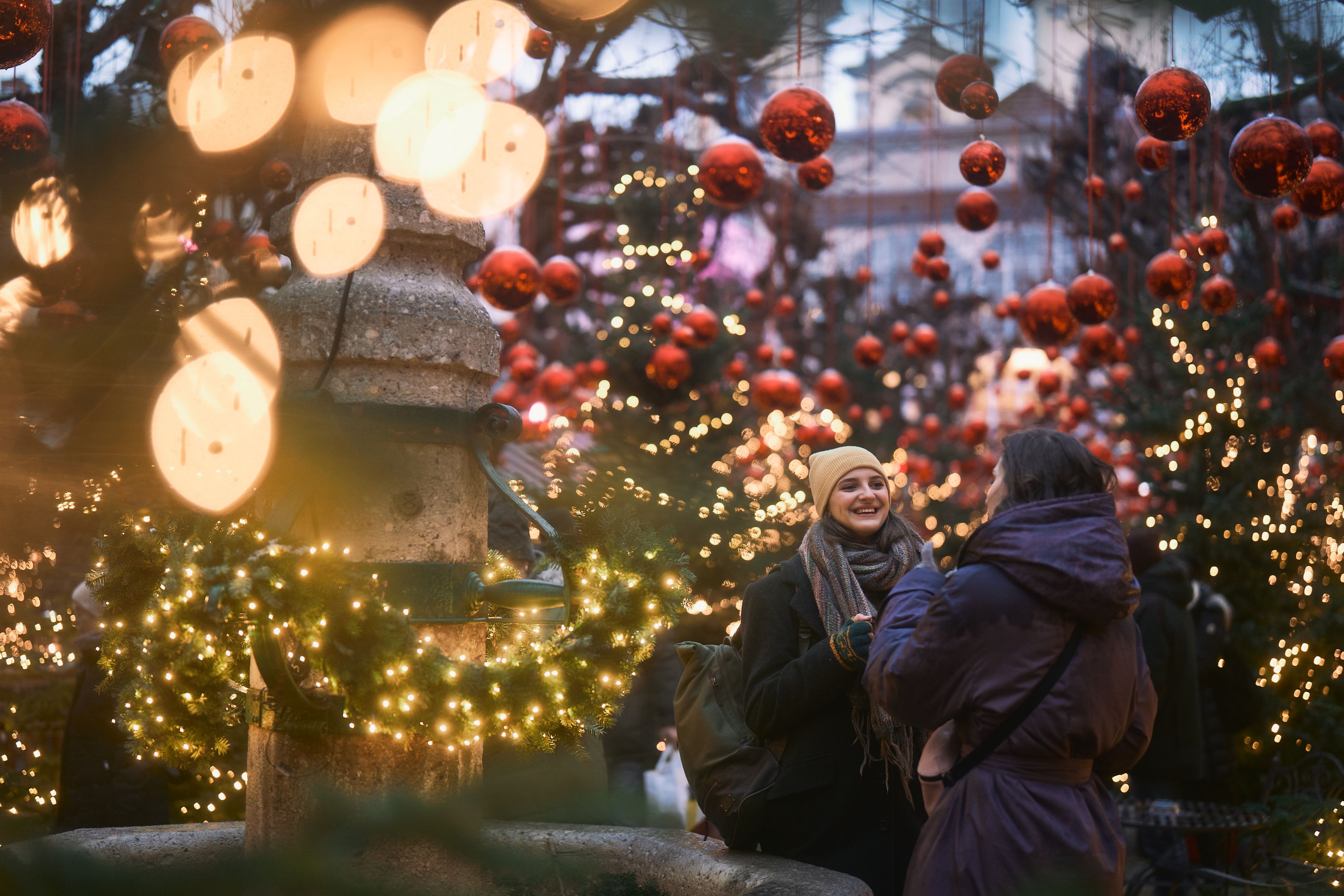 Zwei Menschen unter Lichterketten und roten Kugeln auf einem festlich geschmückten Weihnachtsmarkt.