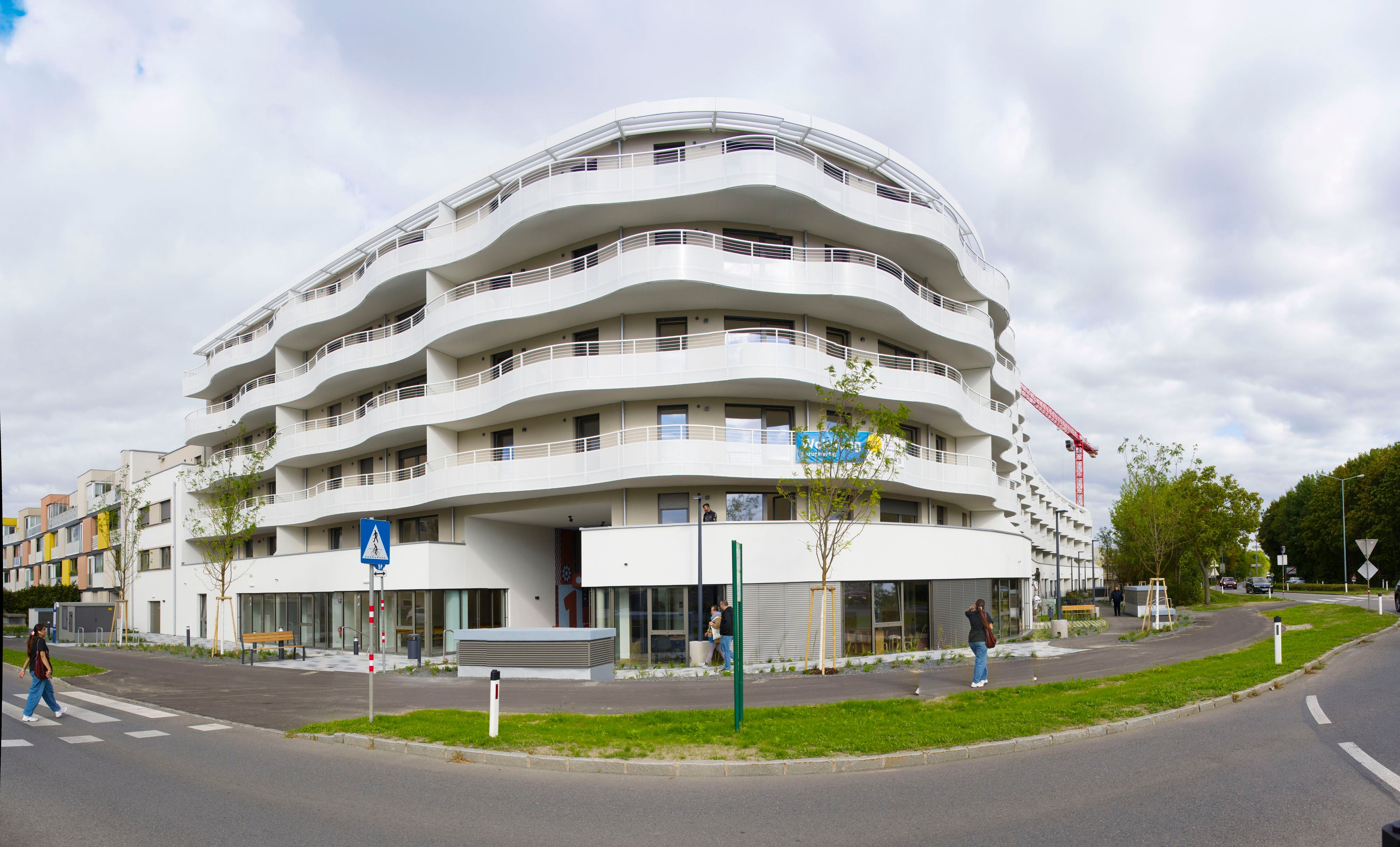 Modernes, weißes Wohngebäude mit geschwungenen Balkonen an einer Straßenecke, Menschen gehen vorbei.