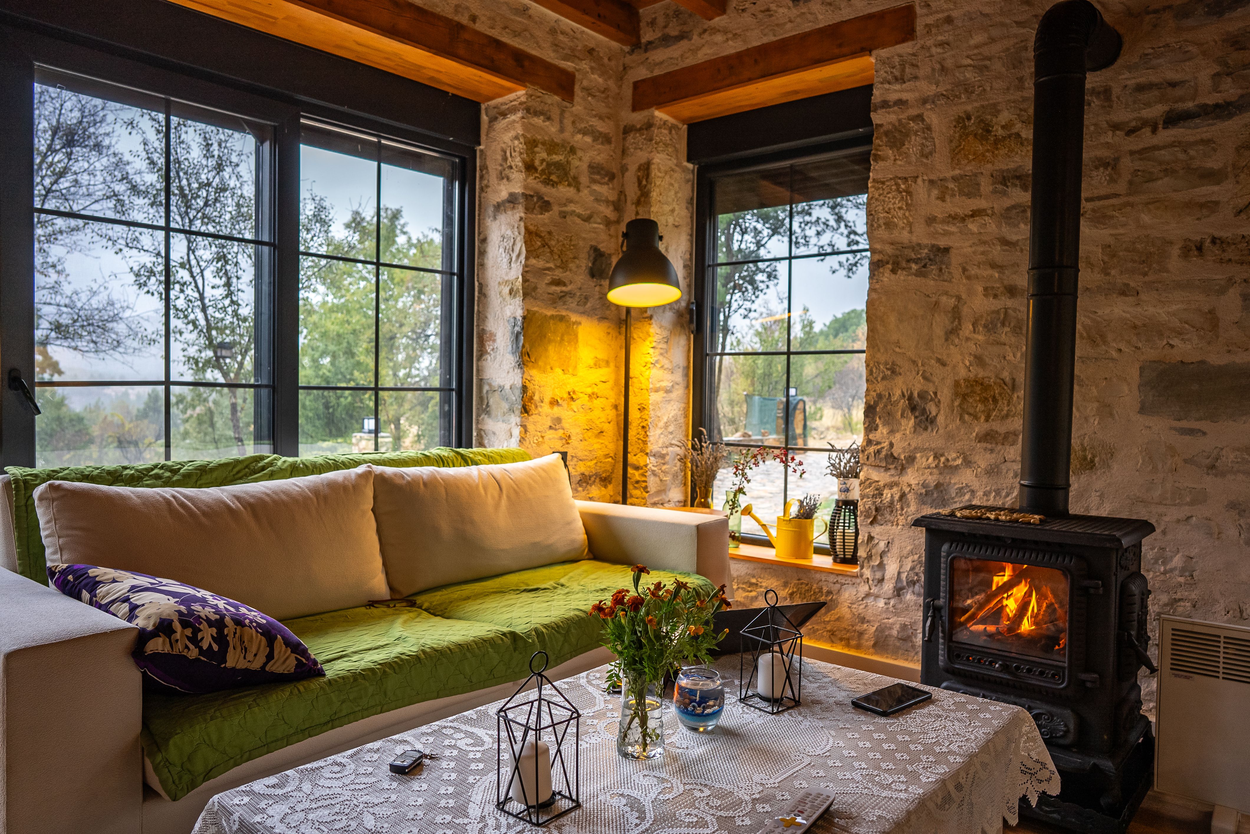 Gemütliches Wohnzimmer mit Sofa, Holztisch, Kaminofen und Blick aus großen Fenstern auf Bäume.