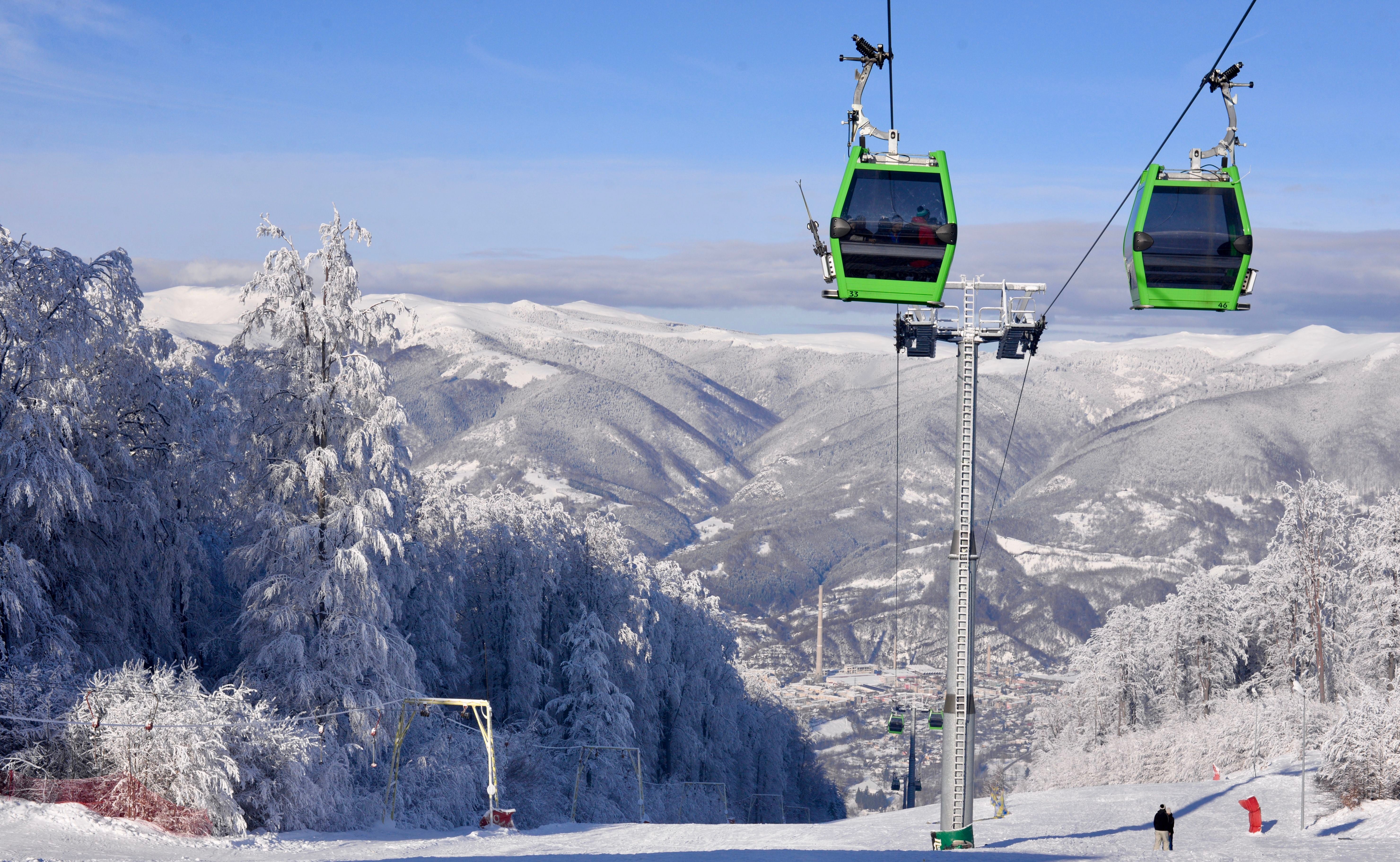 Grüne Gondeln fahren über ein verschneites Skigebiet mit Blick auf Berge und eine Kleinstadt.