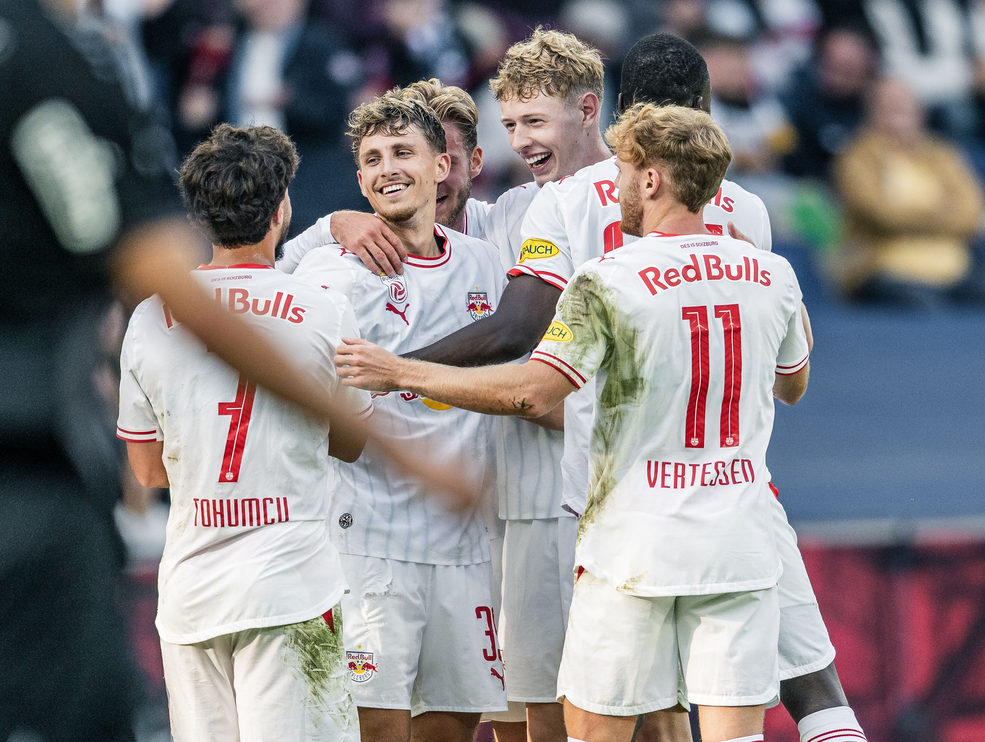 Mehrere Spieler von Red Bull Salzburg in weißen Trikots umarmen sich strahlend auf dem Spielfeld und feiern gemeinsam ein Tor.